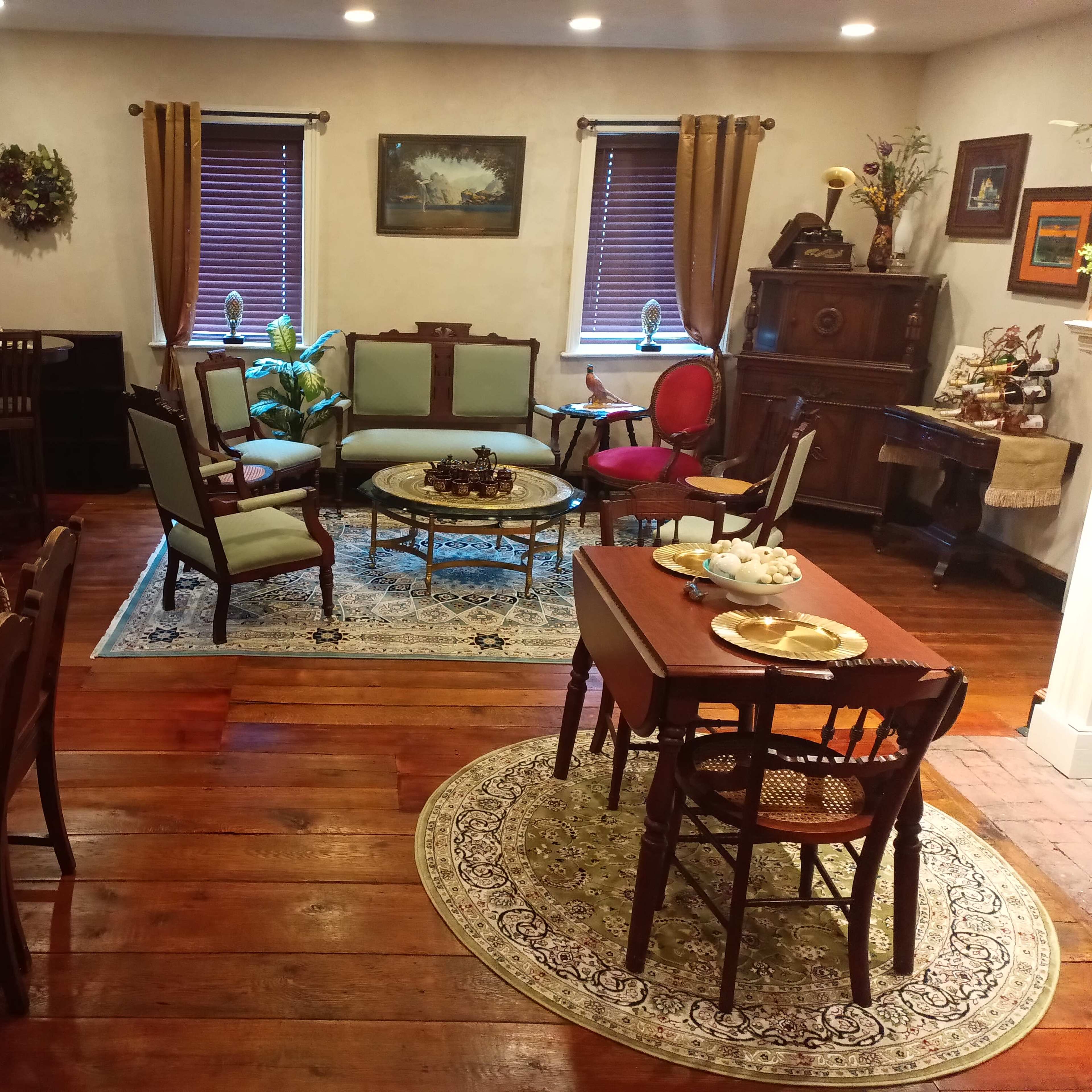 A vintage-style dining and living area with wooden floors, a table set for a meal, and several antique chairs and furniture pieces arranged around the room.