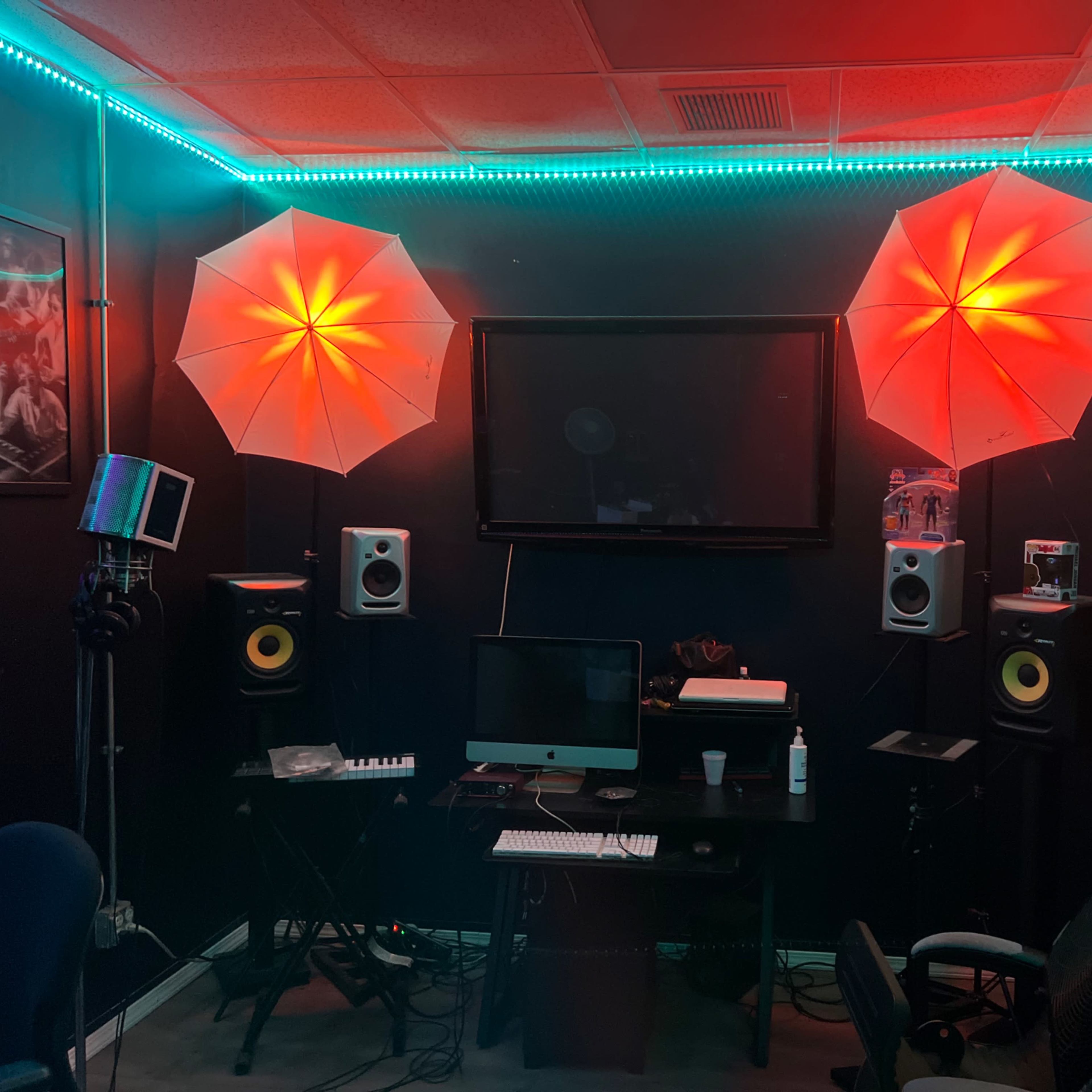 A music studio setup featuring a computer desk, speakers, and red-lit umbrella lights on either side of a wall-mounted screen.