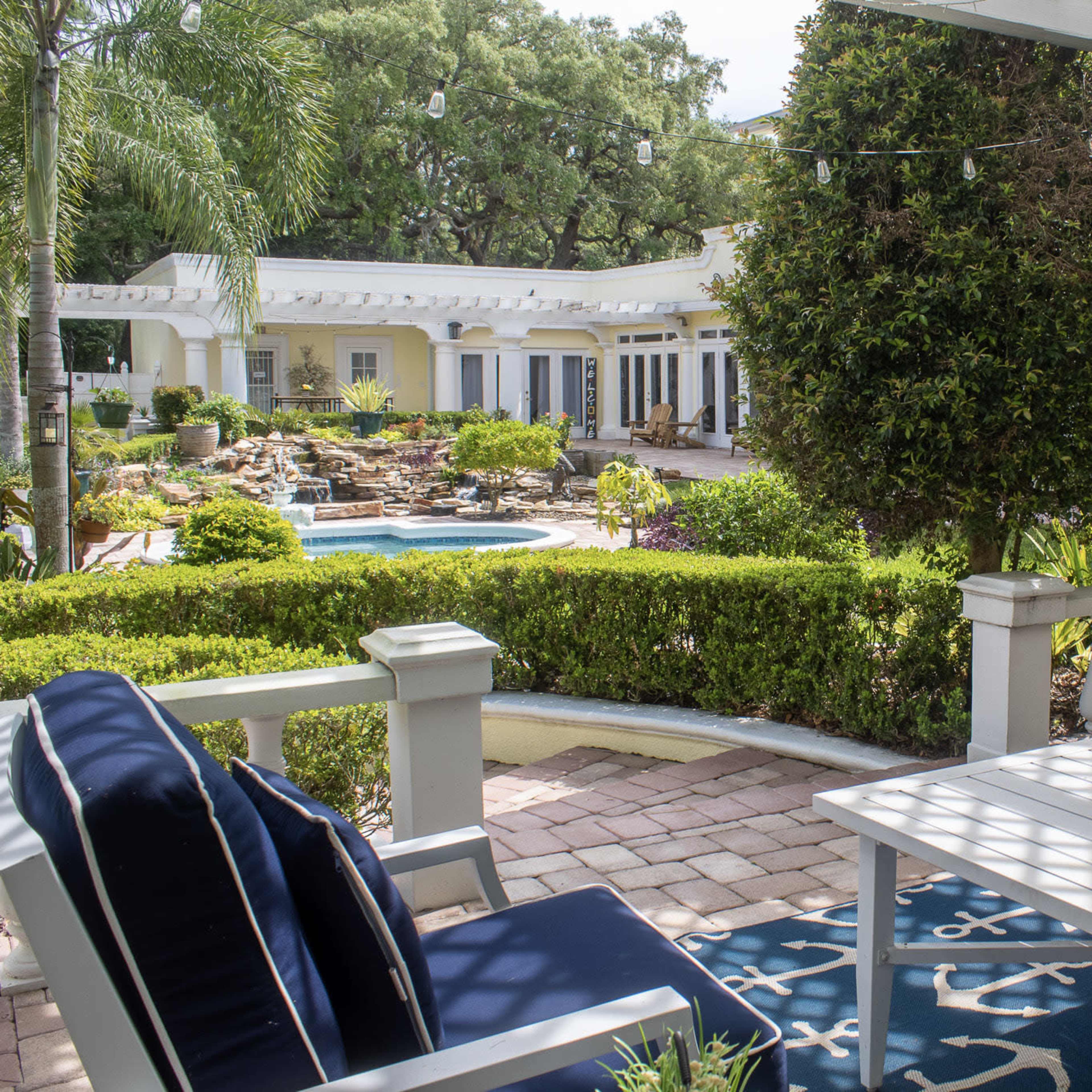 A patio with blue cushioned chairs and a table overlooks a landscaped garden featuring a pool and neatly trimmed hedges.
