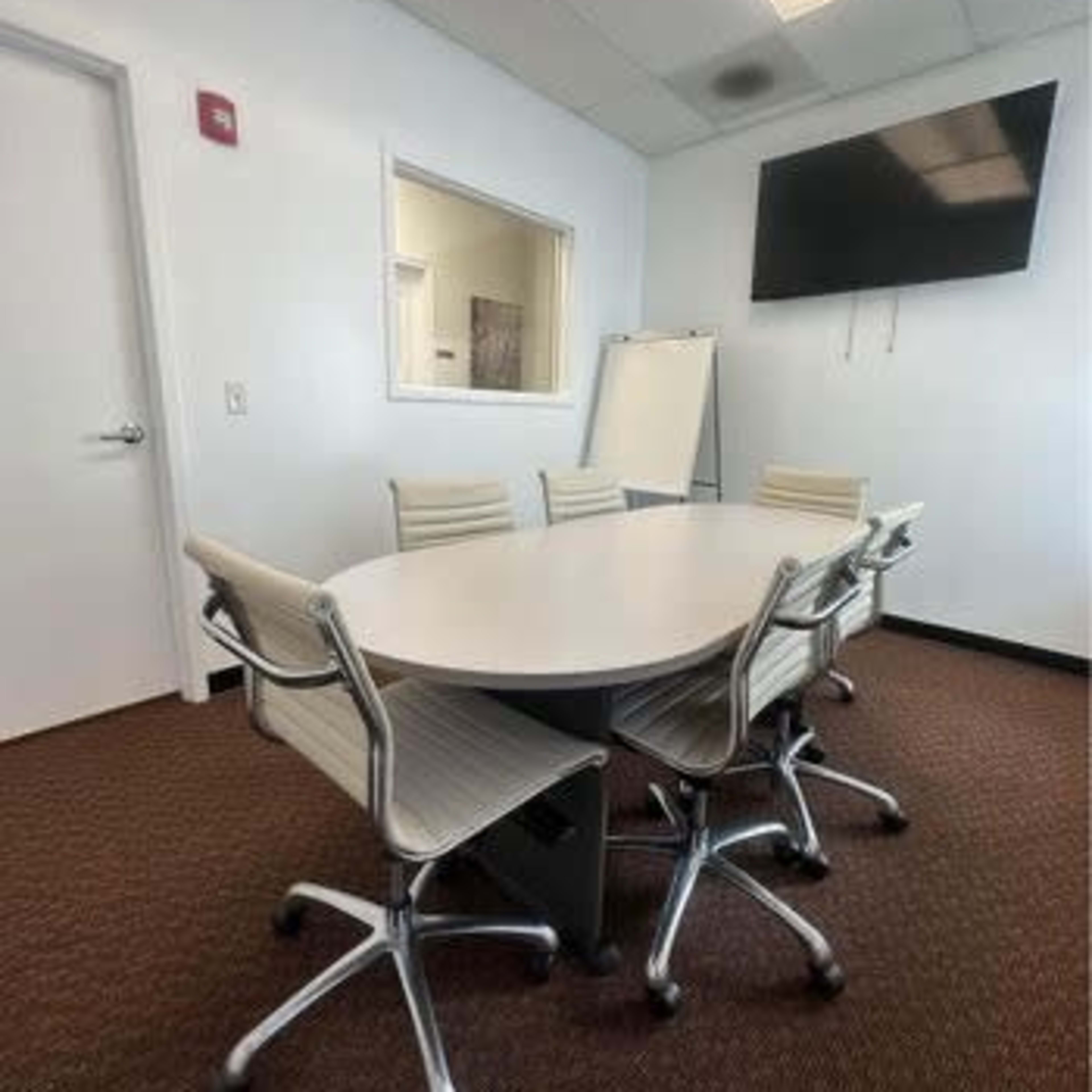 A conference room with a large oval table surrounded by six modern chairs and a wall-mounted television.