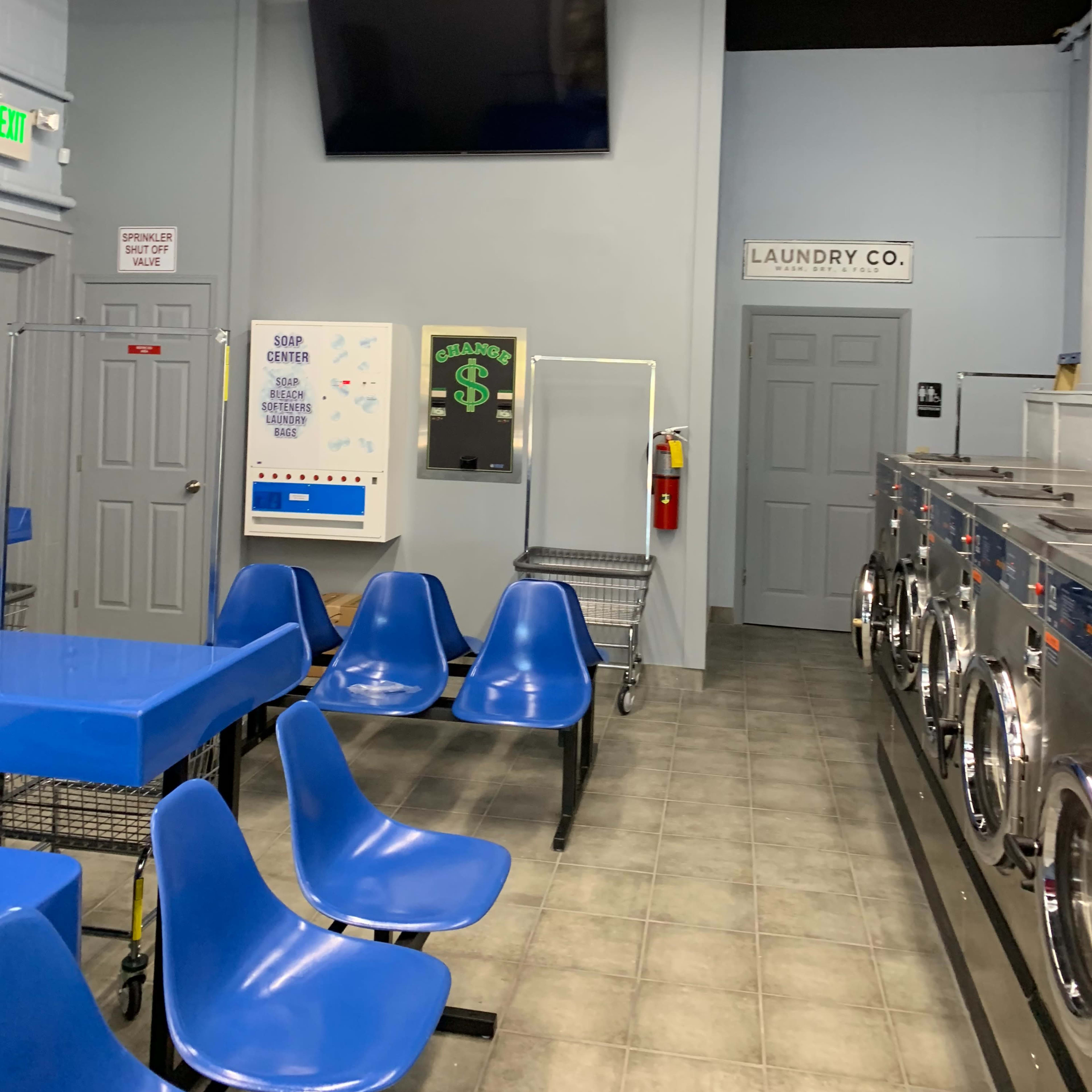 The interior of a laundry facility featuring blue plastic chairs, a wall-mounted television, and rows of washing machines.