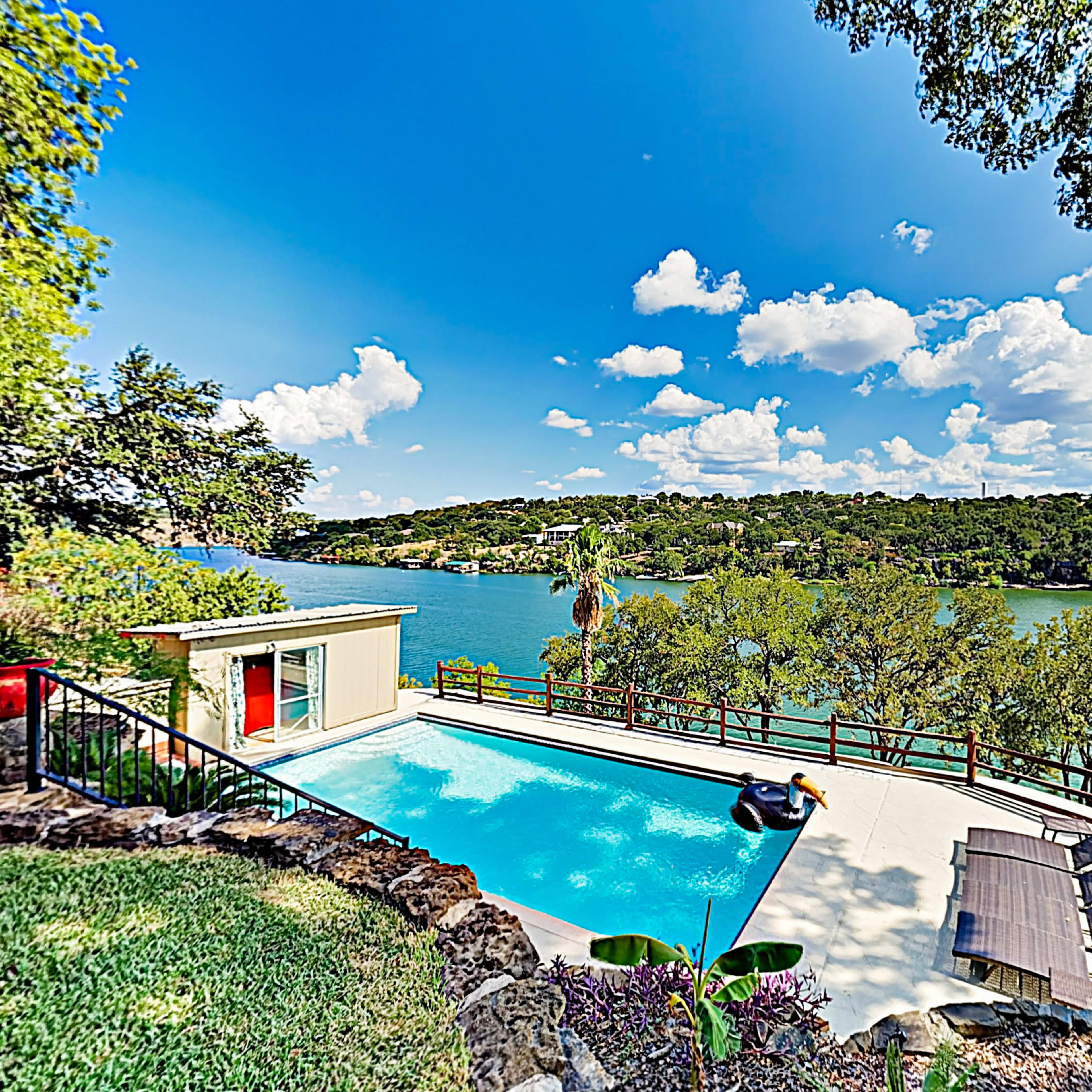 A swimming pool overlooks a river surrounded by trees and a clear blue sky filled with clouds.