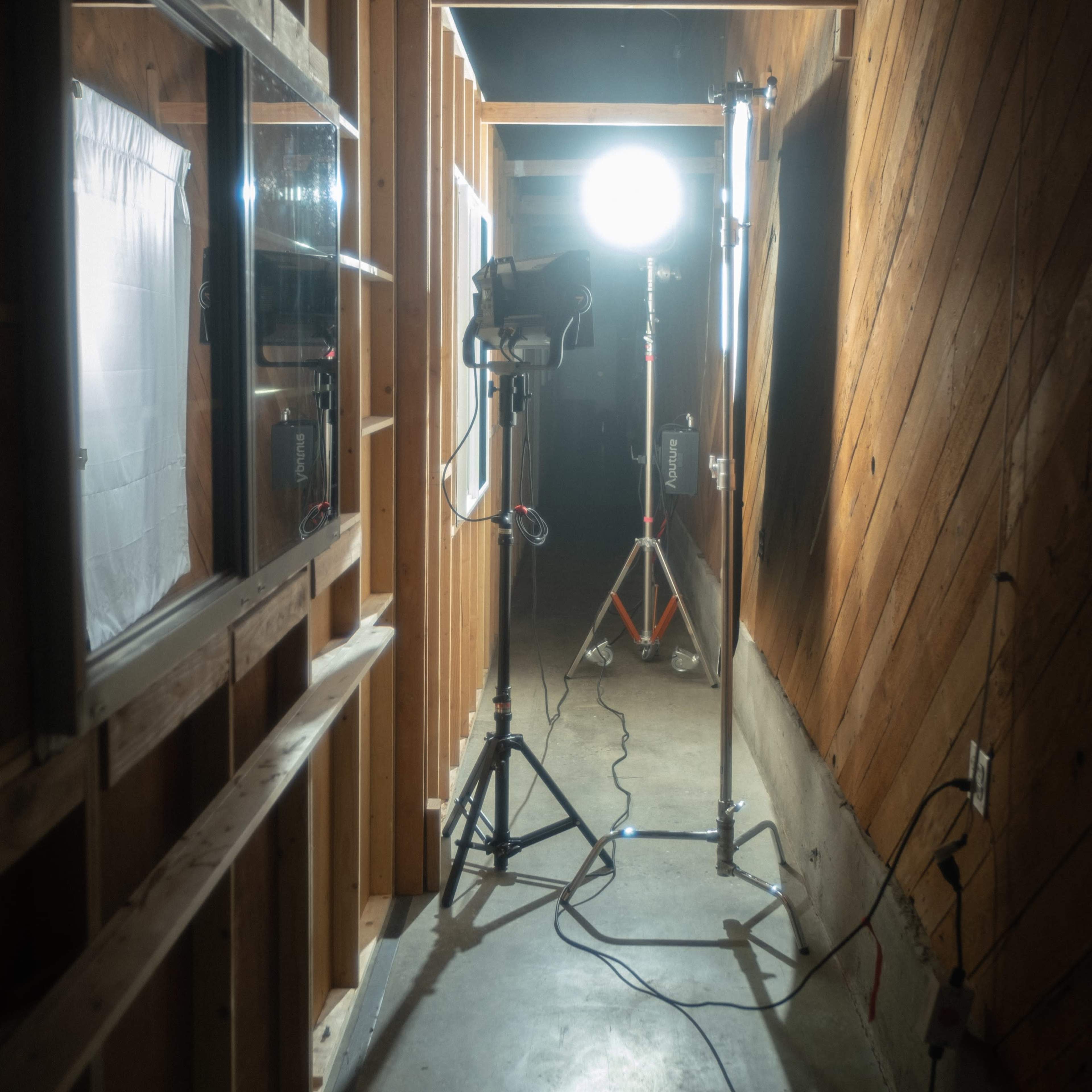 A narrow hallway with wooden walls features a bright overhead light and photographic lighting equipment set up on stands.