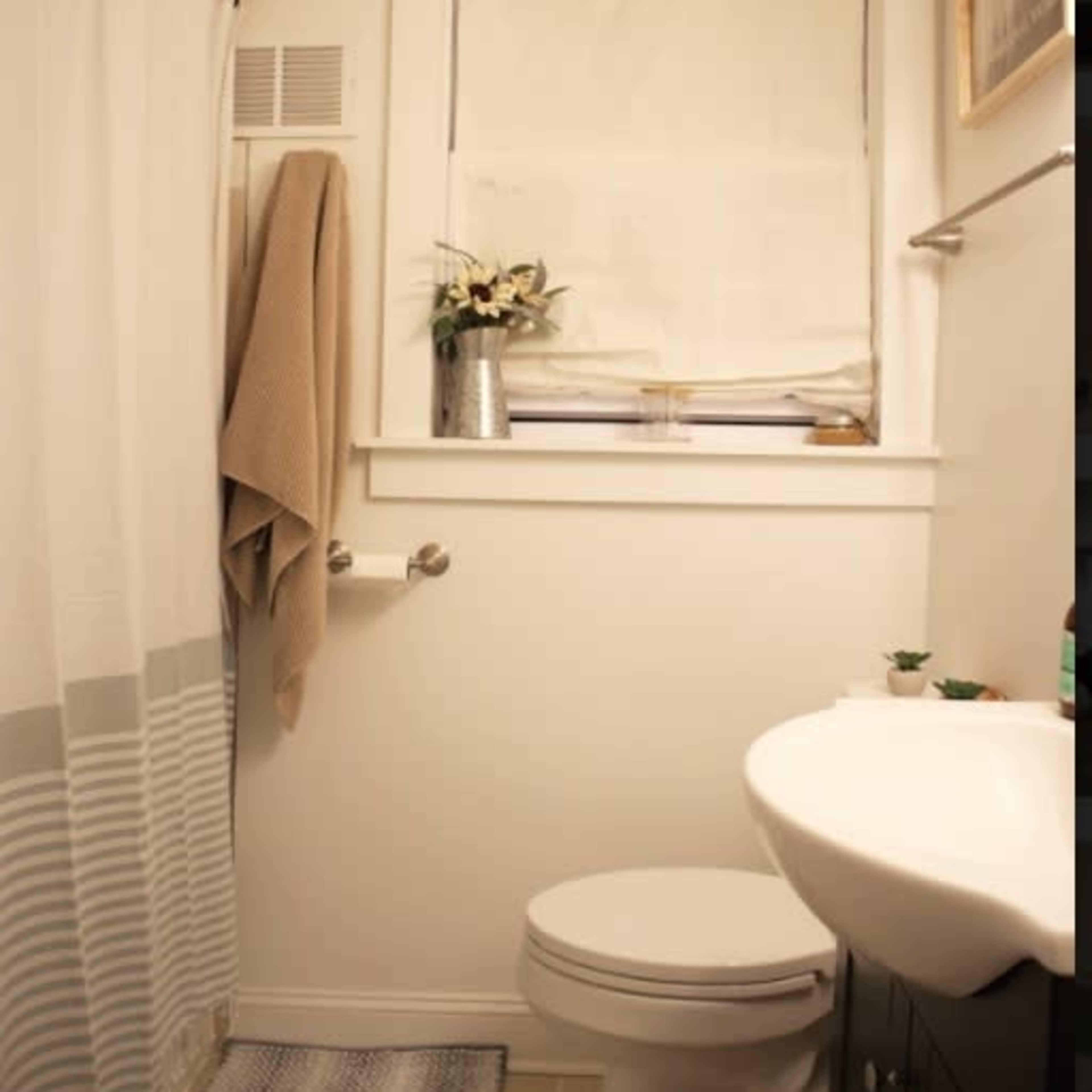 A small bathroom featuring a toilet, sink, window with blinds, a shower curtain, and a decorative towel hung on the wall.
