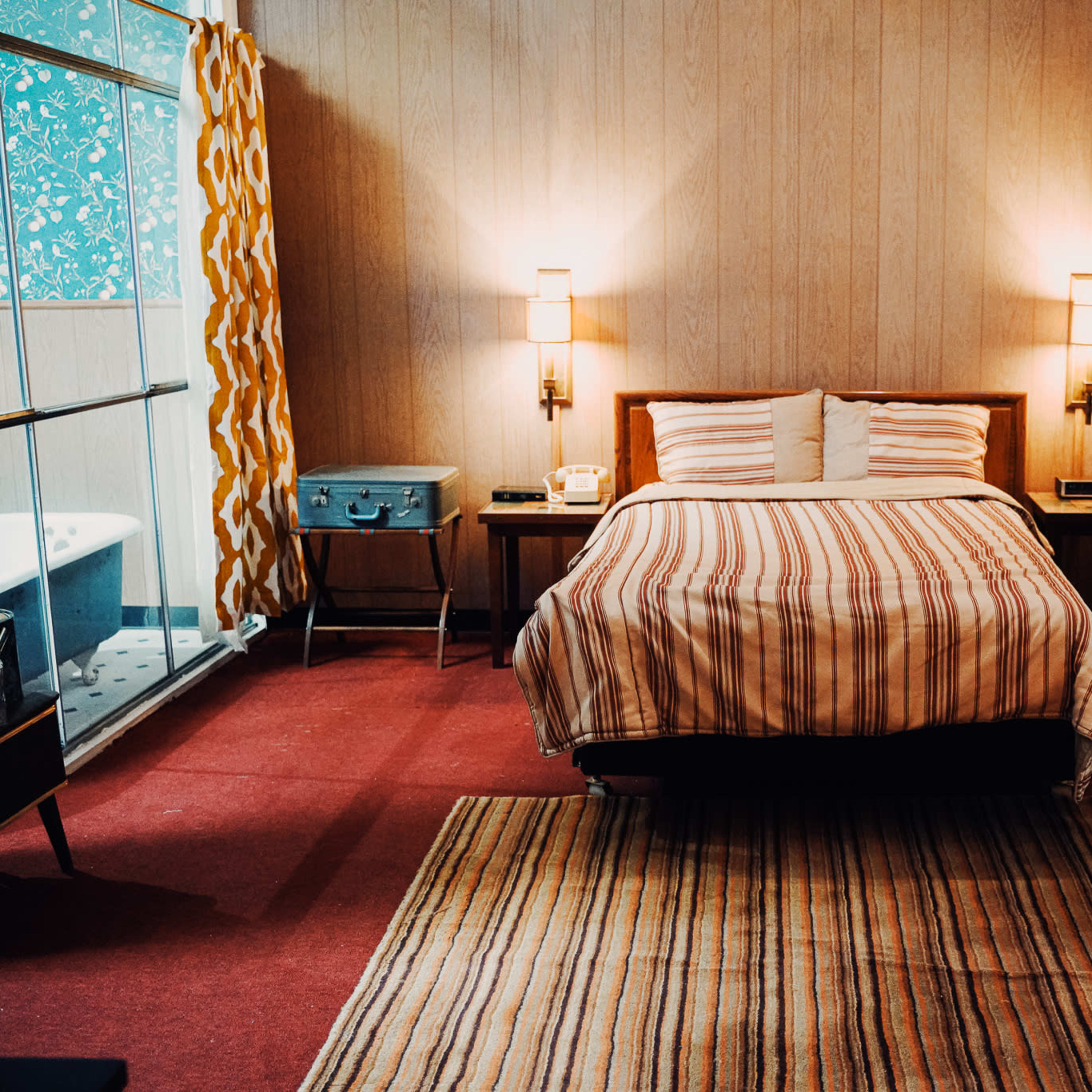 A retro-style hotel room featuring a bed with striped bedding, a patterned carpet, and a vintage television placed next to a wooden nightstand.
