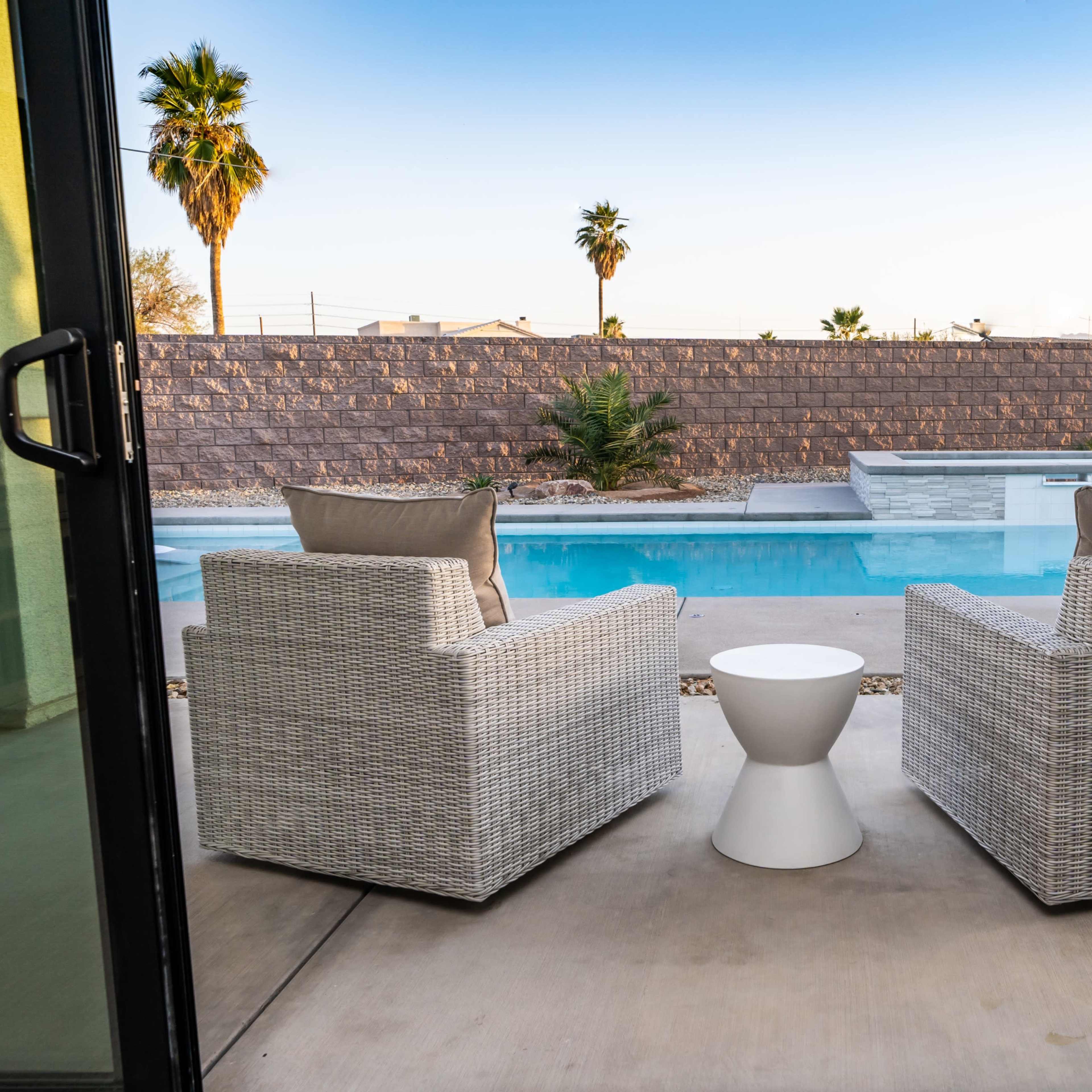 A pair of white wicker chairs and a small round table face a swimming pool surrounded by palm trees and a stone wall.
