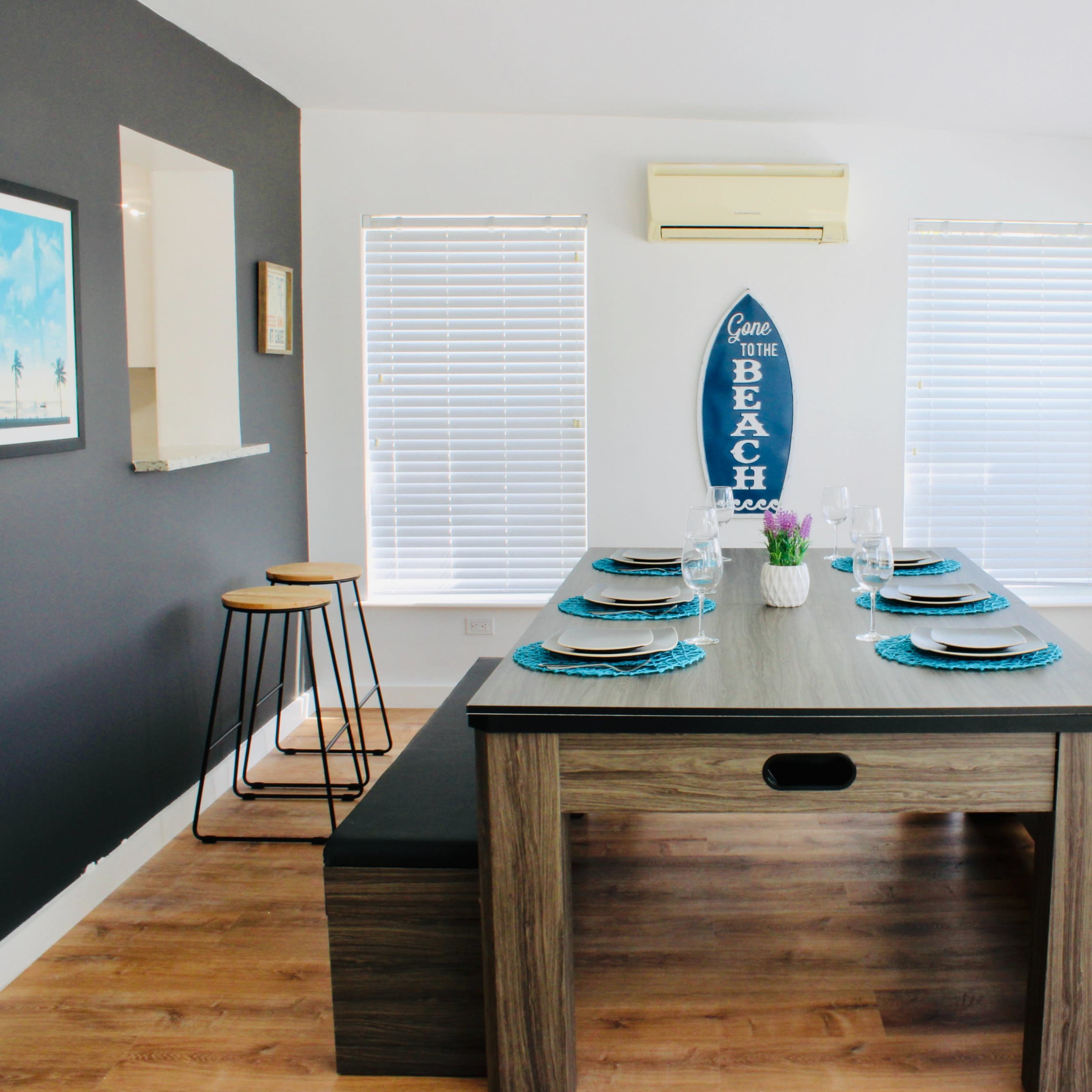 A dining area features a large wooden table with a surfboard-themed wall decoration and windows with blinds.