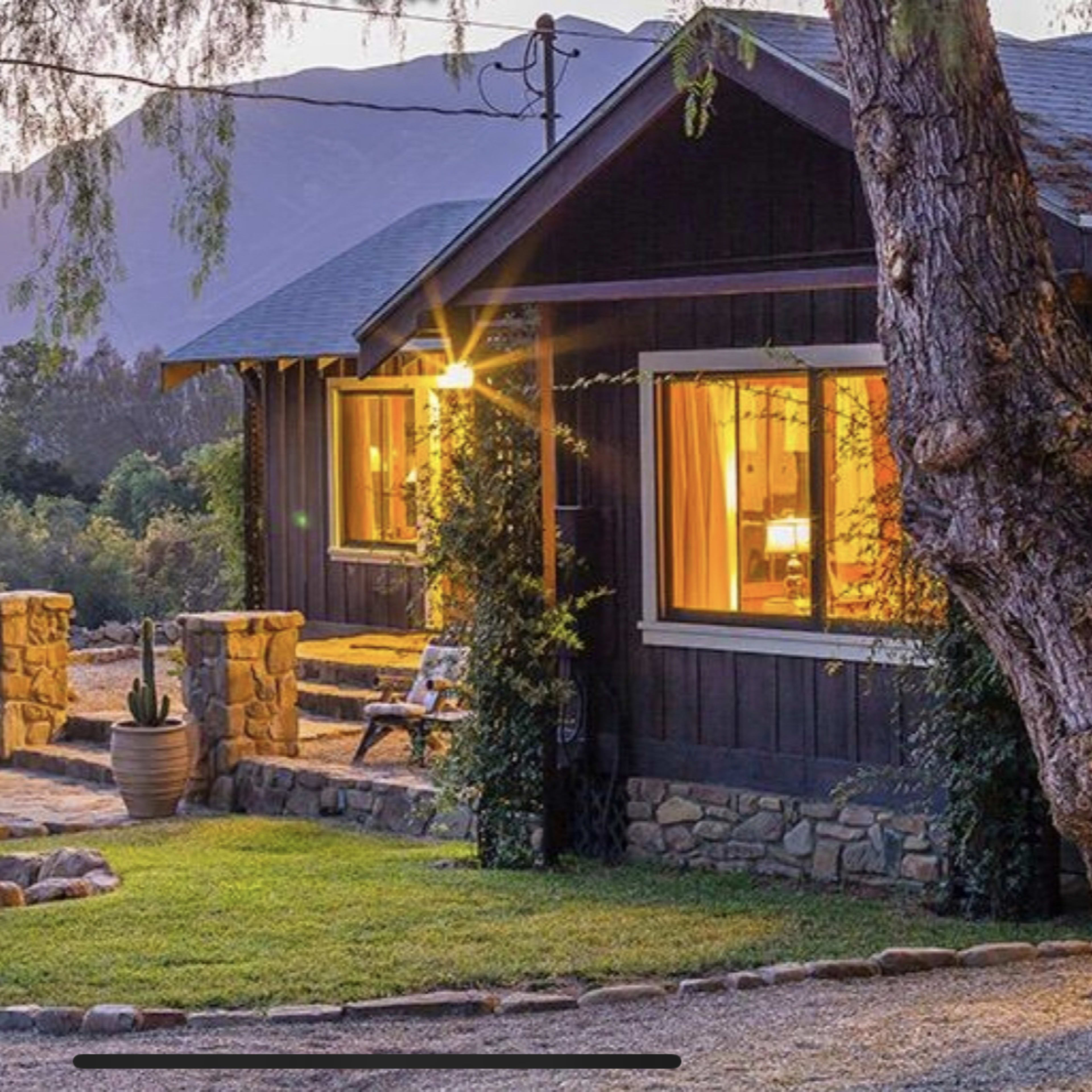 A quaint wooden cottage with warm lights visible through the windows is surrounded by a gravel pathway and a landscaped yard.