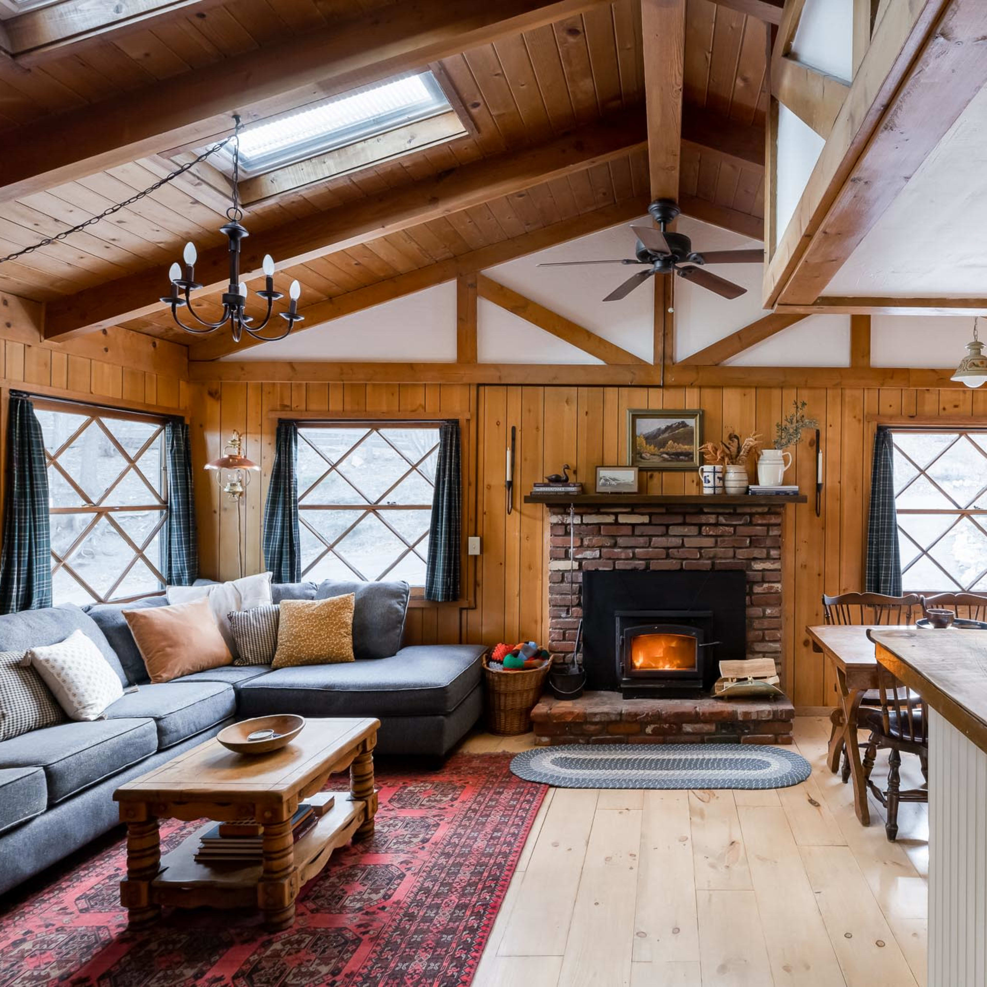 A cozy cabin interior featuring a living area with a brick fireplace, a sectional sofa, and a dining table near a kitchen with rustic decor.