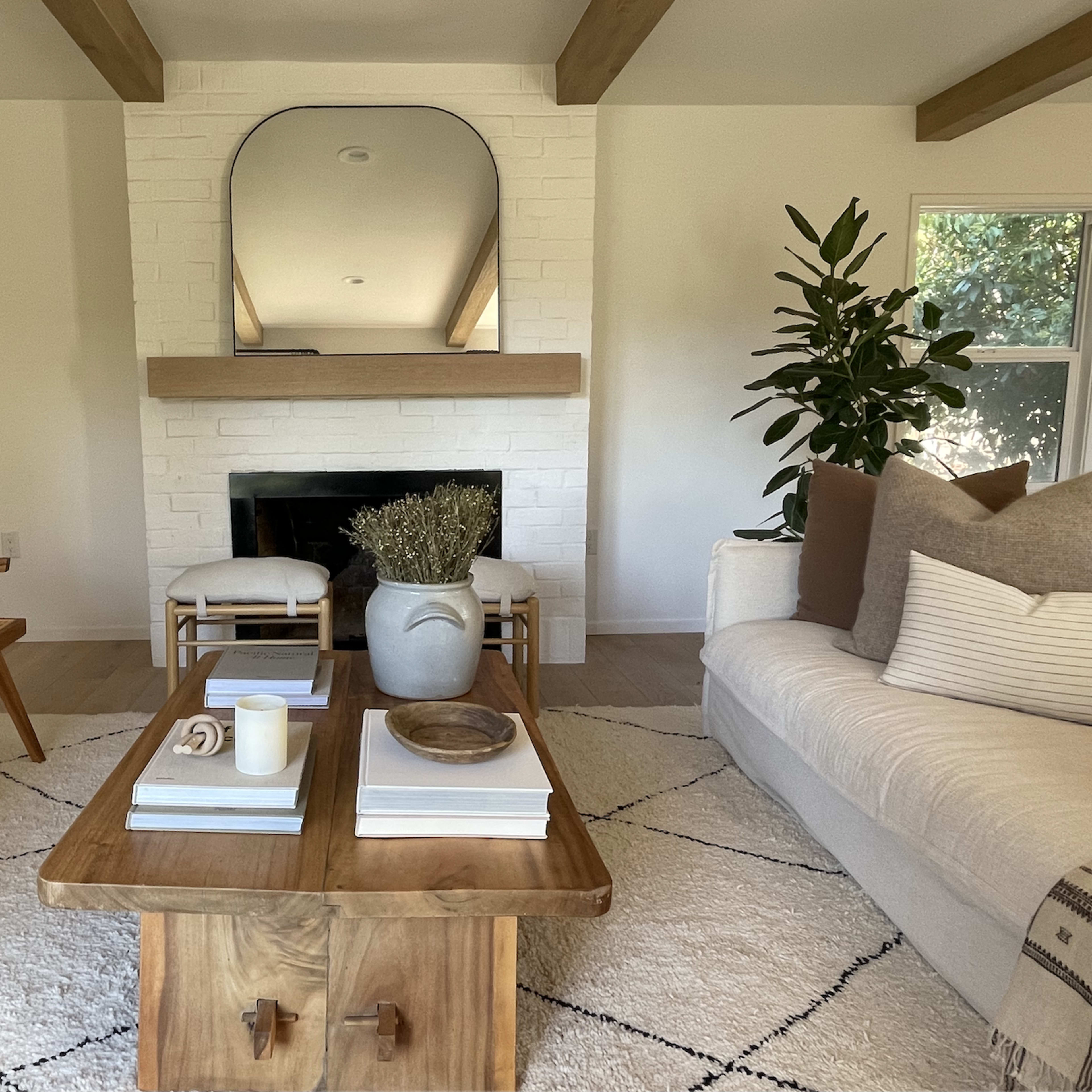 A living room featuring a white sofa, a wooden coffee table, and a minimalist fireplace with a large mirror above it surrounded by plants.
