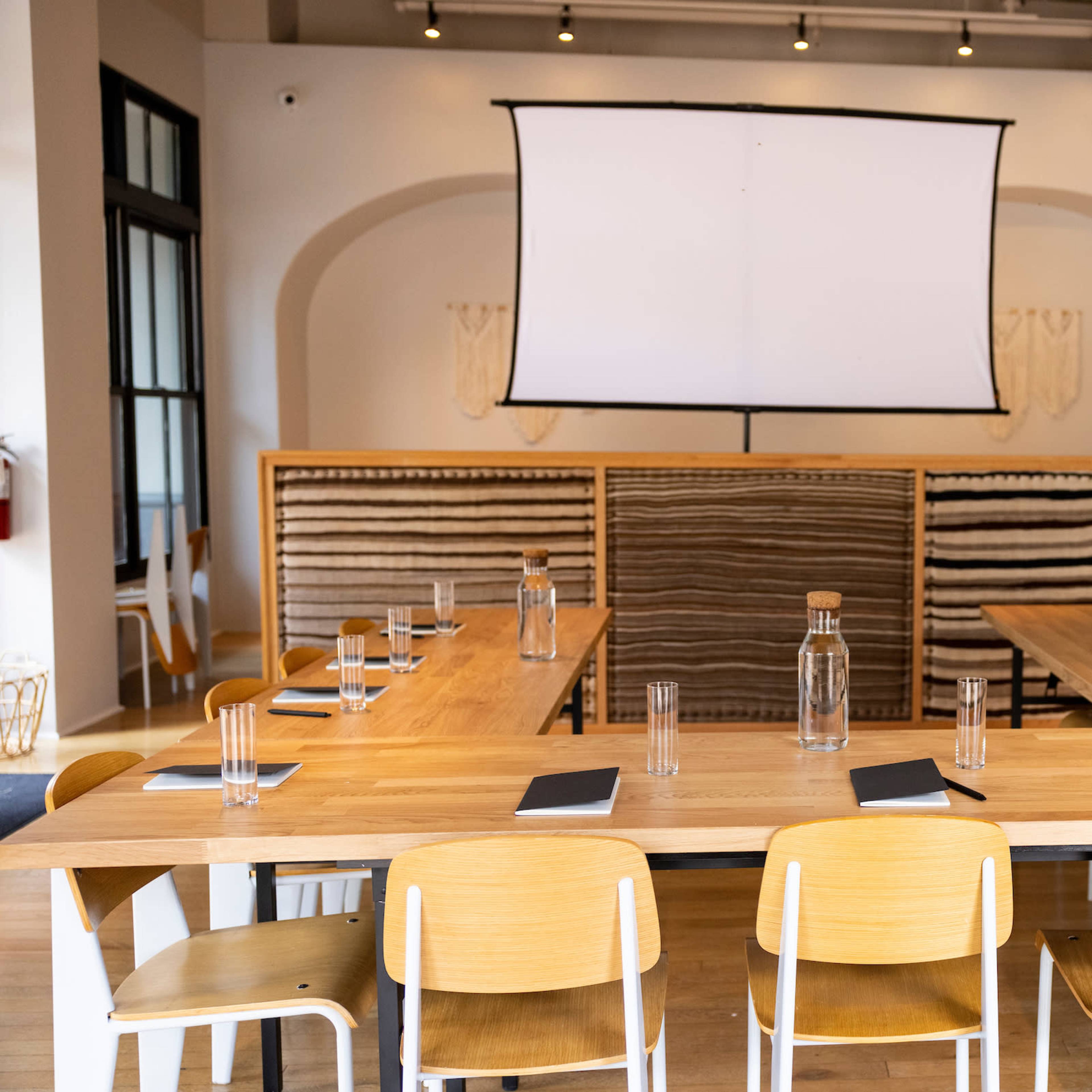 A spacious meeting room features two large rectangular tables arranged in an "H" shape, surrounded by chairs, with a blank projector screen in the center.