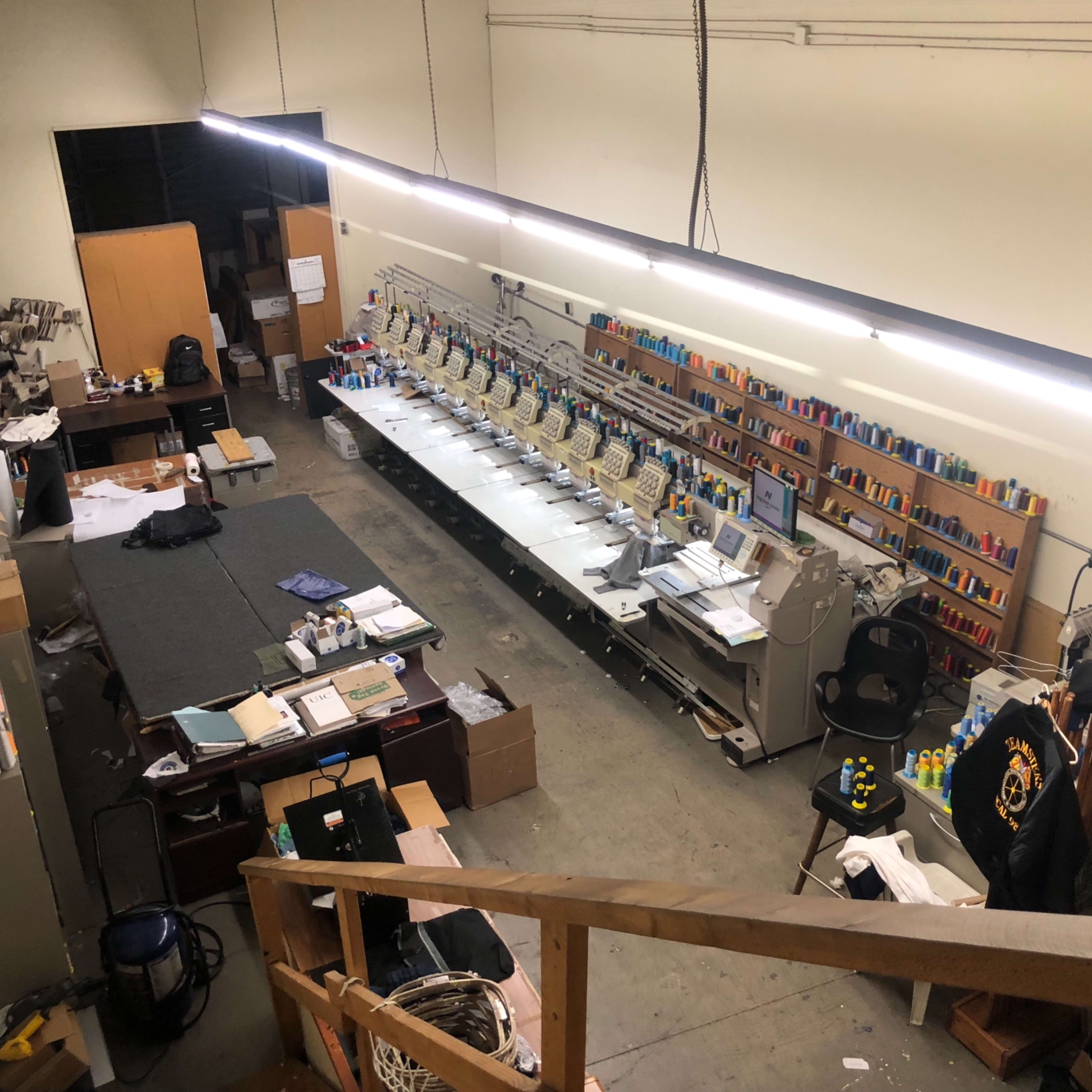 A spacious workshop with a row of sewing machines, various colorful spools of thread on shelves, and scattered materials and equipment around the floor.