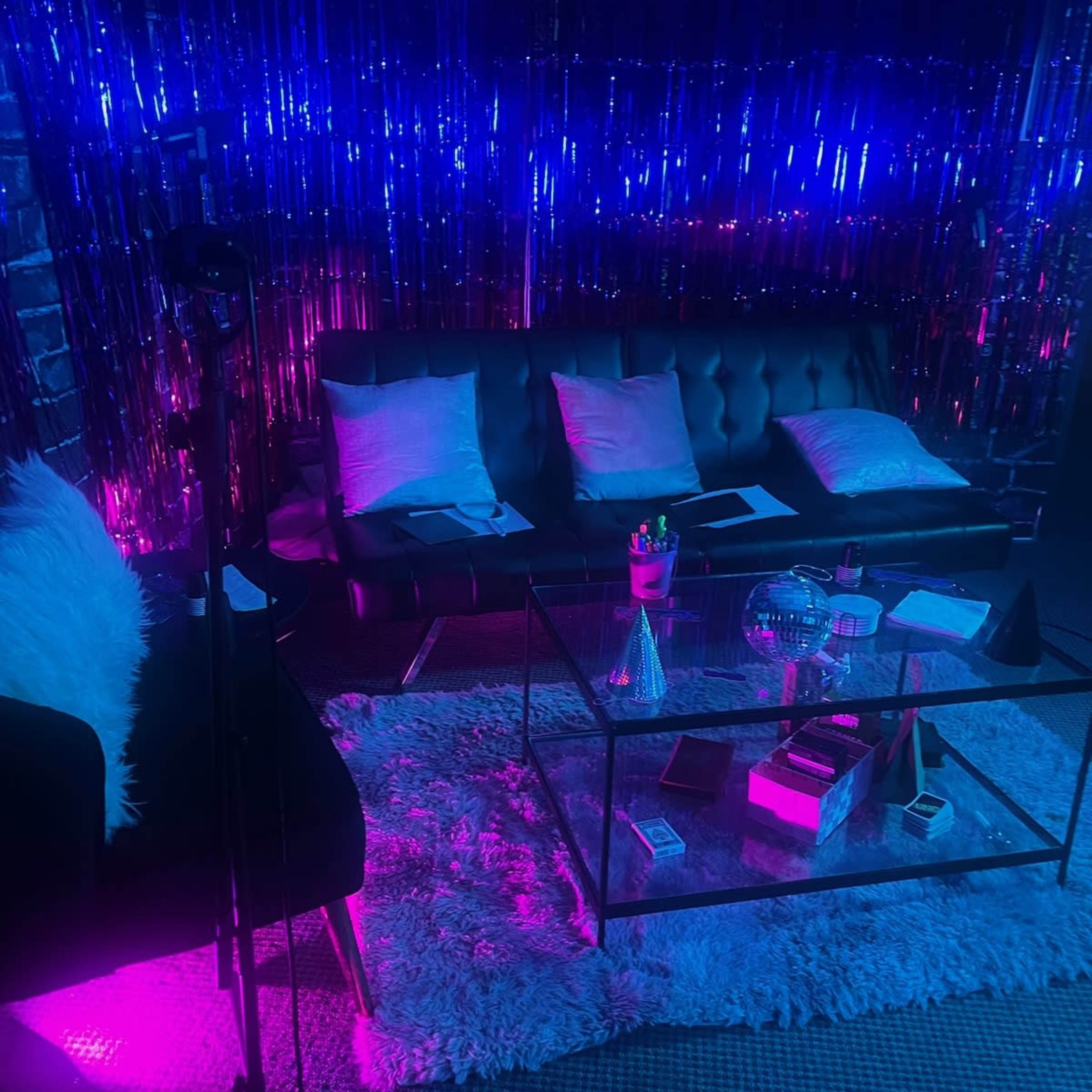 The scene features a modern lounge area with a black leather sofa, a glass coffee table, and colorful lighting effects created by shiny decorations in the background.