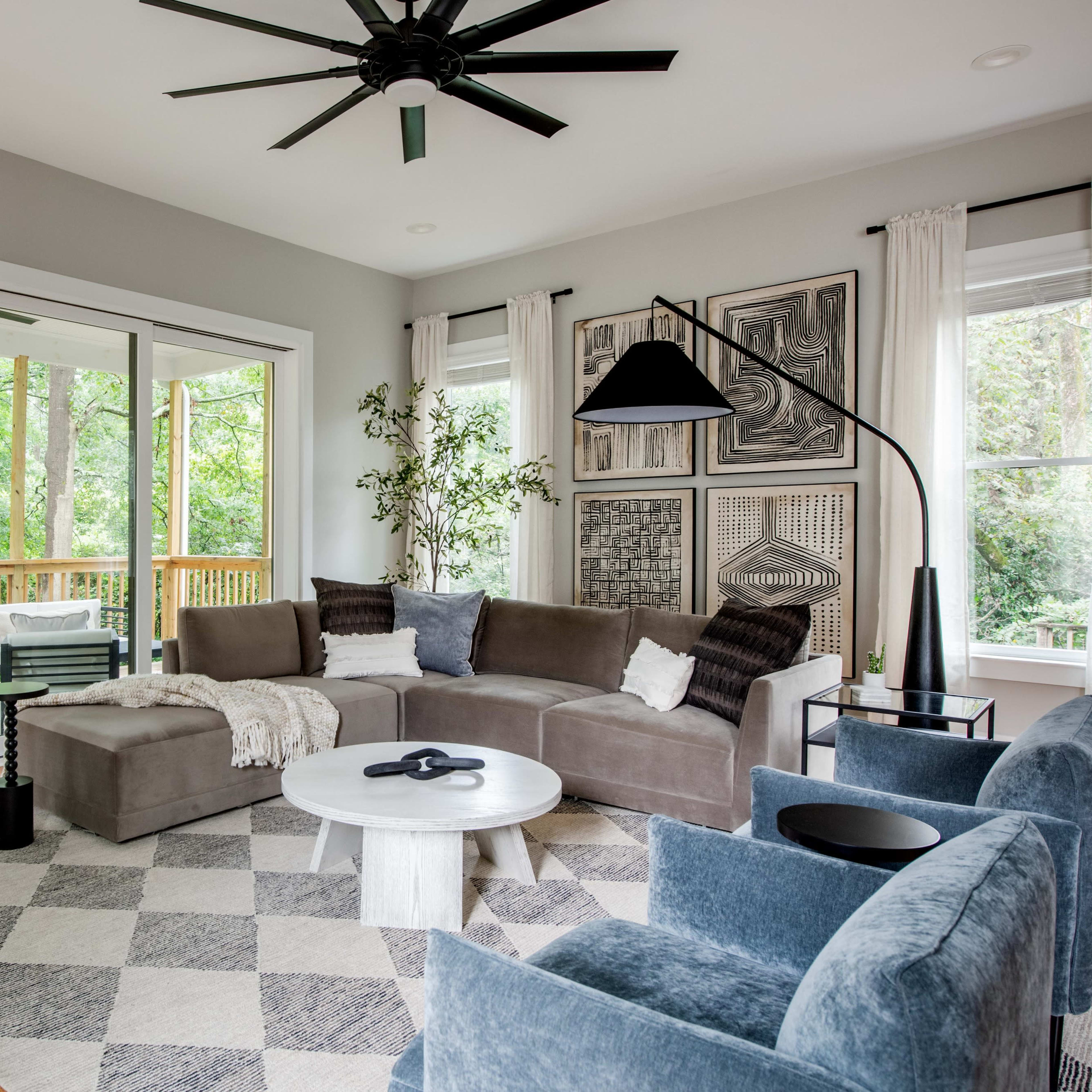 A modern living room with a gray sectional sofa, blue accent chairs, a round coffee table, and large windows overlooking a deck surrounded by trees.