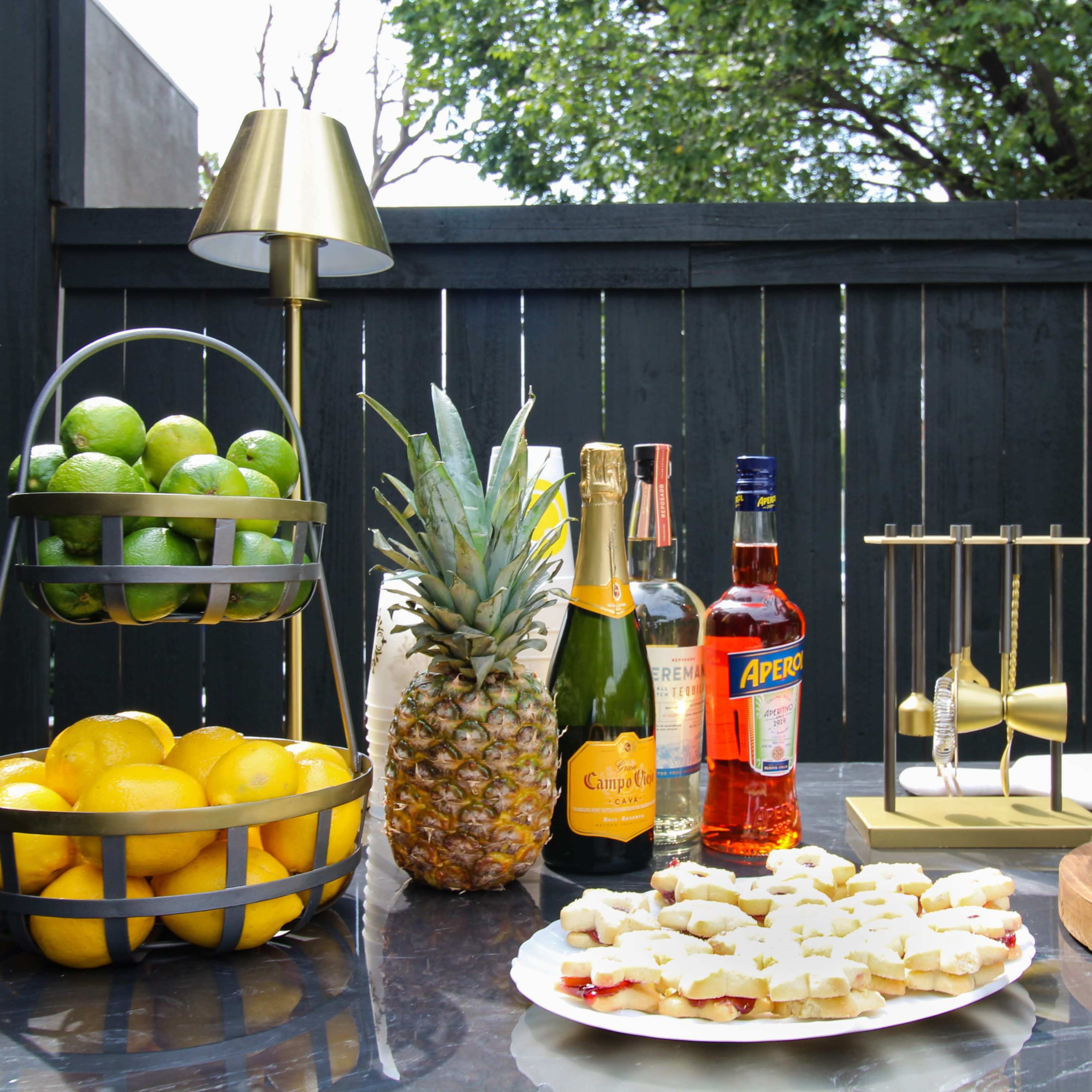 A table is set outdoors featuring a tiered fruit basket with limes and lemons, a pineapple, bottles of champagne, Aperol, and a cocktail shaker, alongside a plate of snacks.