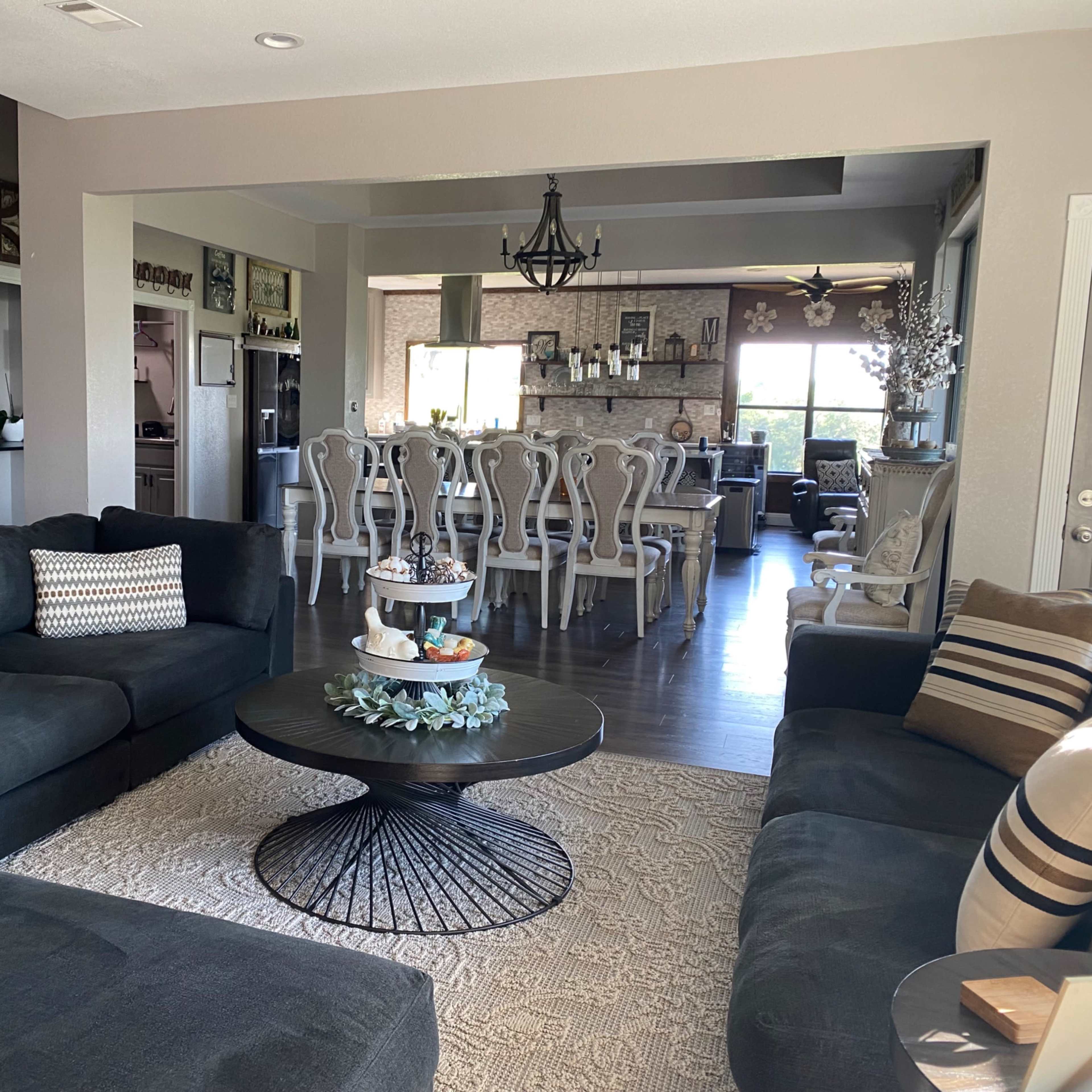 A spacious living room with dark sectional sofas, a round coffee table, and a decorative area rug, leading into a dining area with white chairs and a large table.