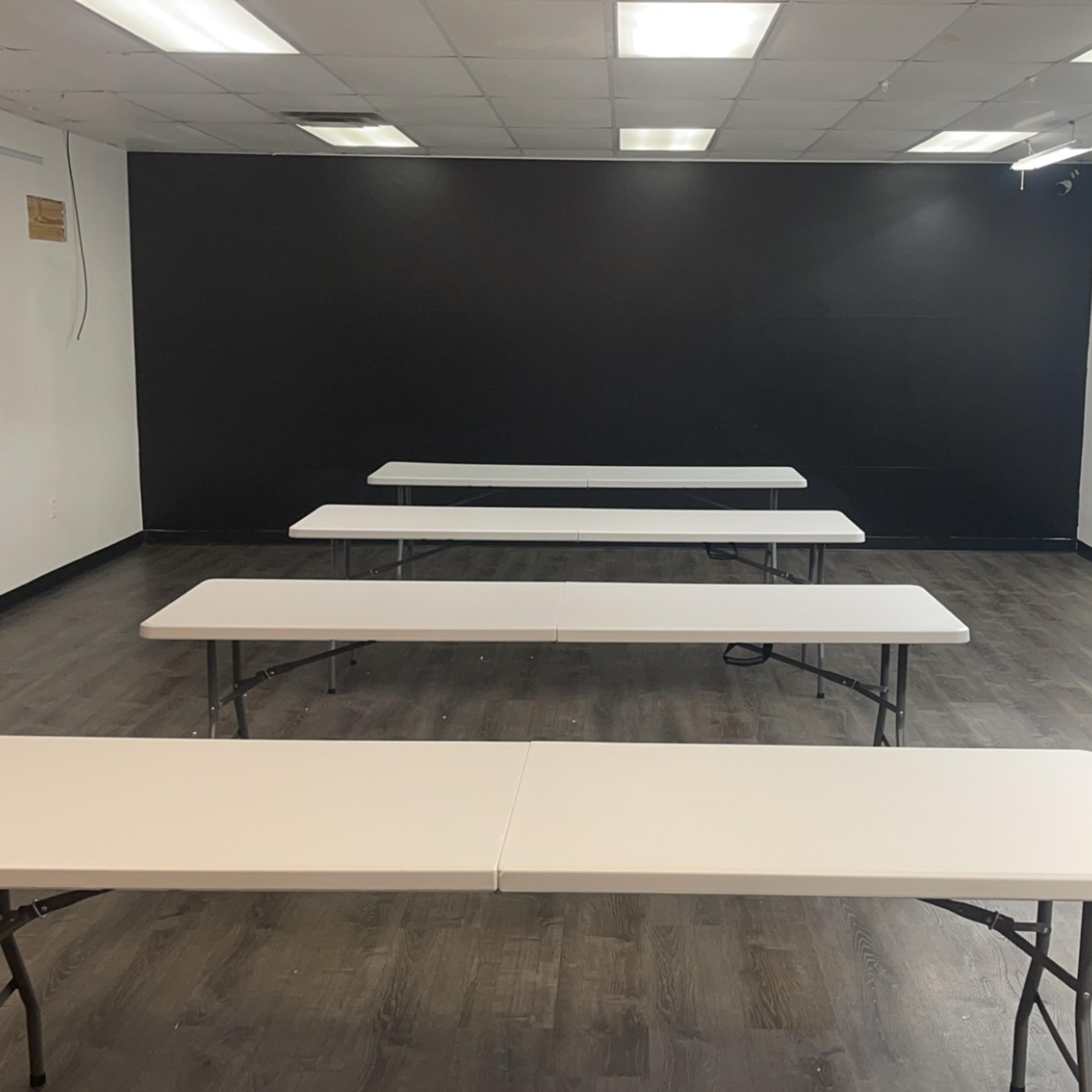 A room with several folding tables arranged in a row against a black wall, with a sink and a utility area visible in the background.