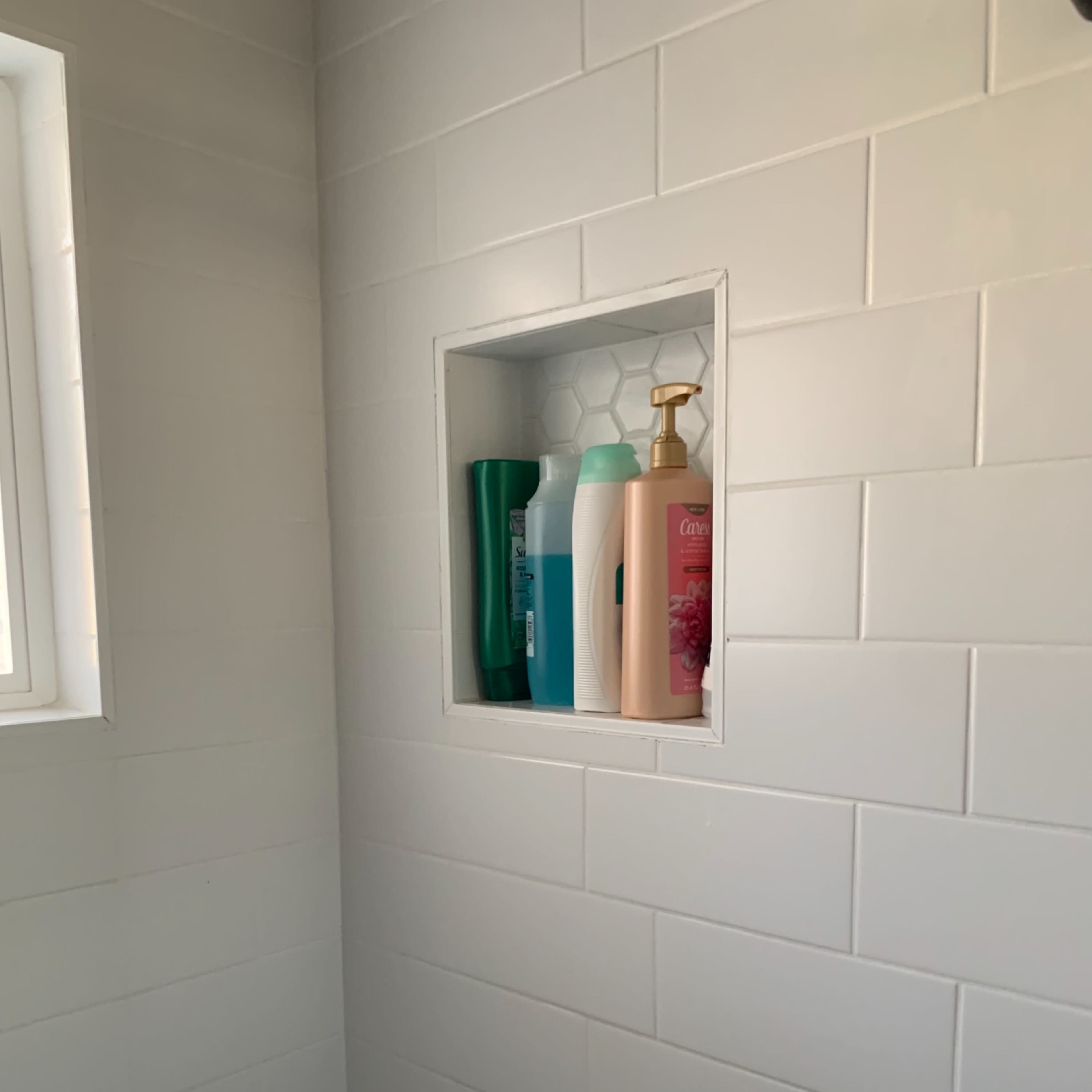 A corner of a shower features a built-in niche holding various bottles of shampoo and body wash against white tiled walls.