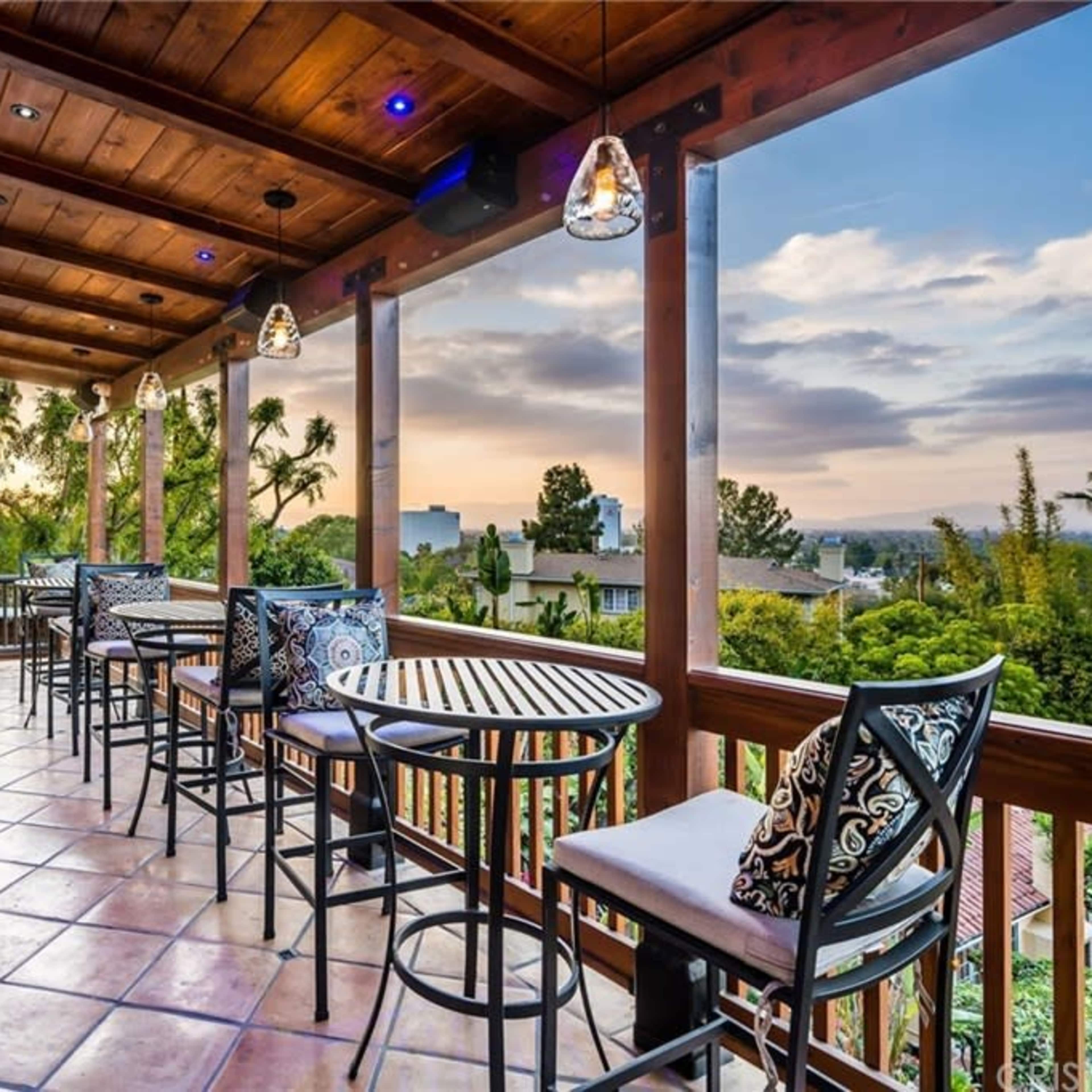 A spacious balcony with bar seating and pendant lighting overlooks a landscape of trees and distant buildings under a cloudy sky.