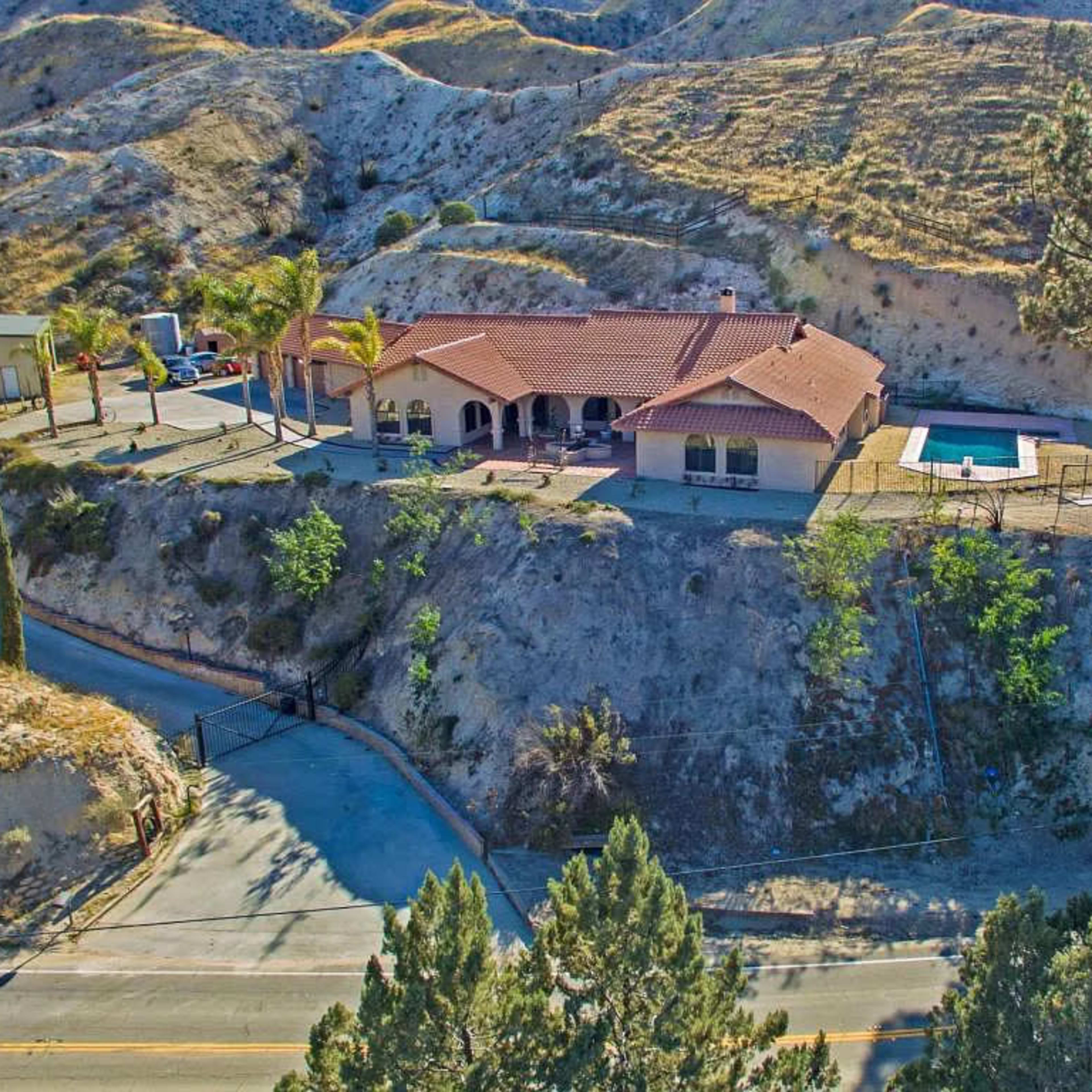 A single-story house on a hillside with a swimming pool, surrounded by trees and a winding road below.