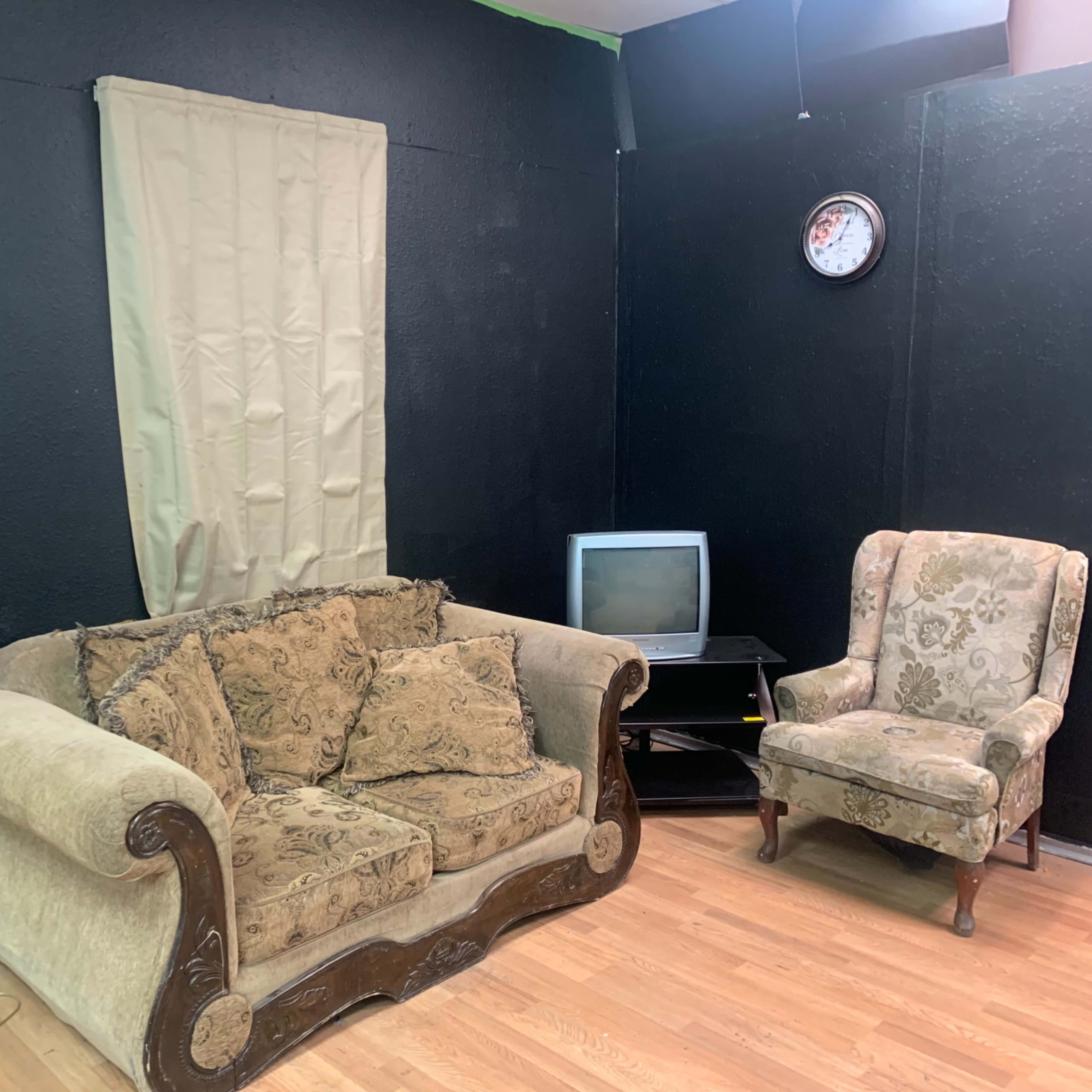 A small living area with a lamp, a patterned armchair, a vintage-style couch with cushions, a television on a stand, and a clock on the wall, all set against a dark wall with a curtain.