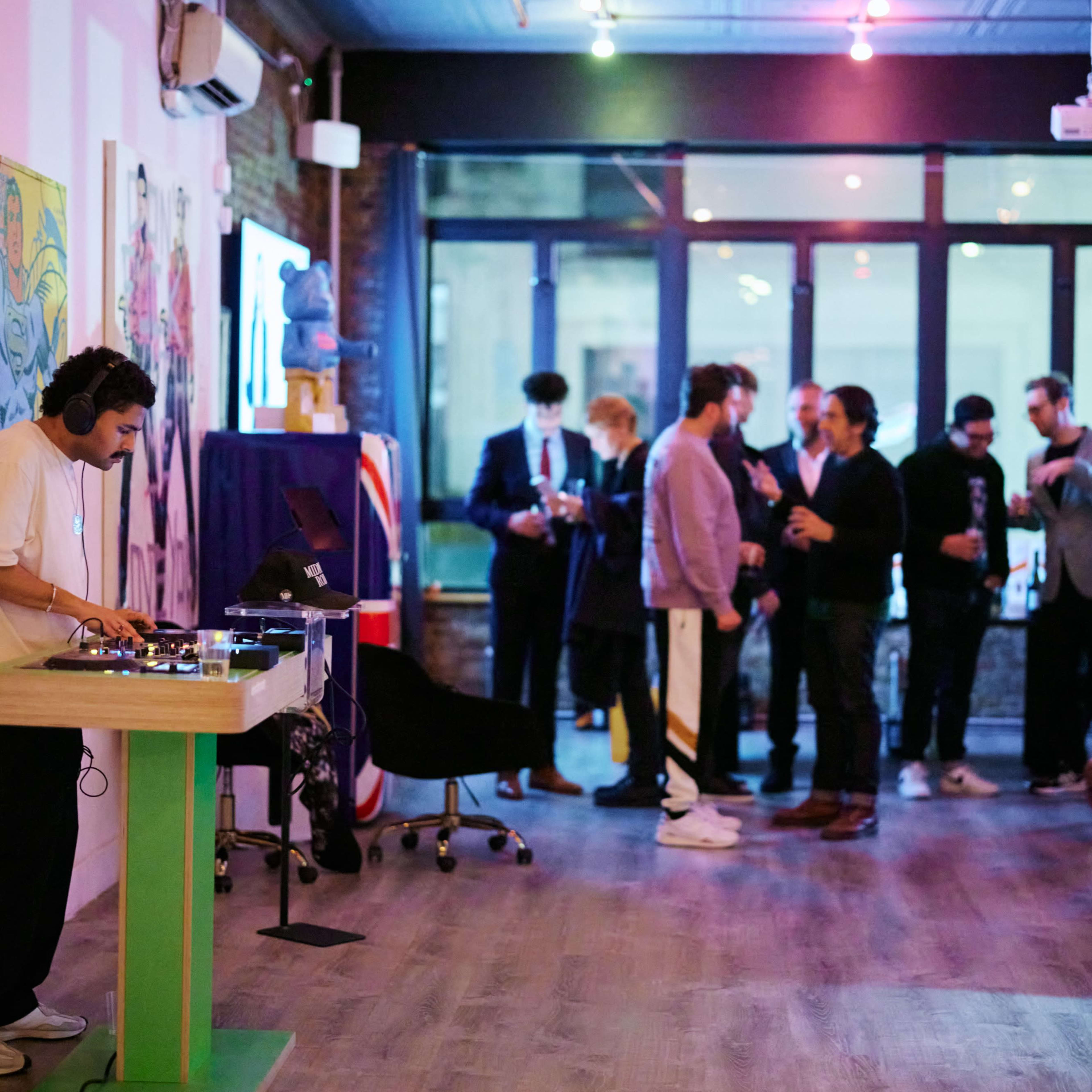 A DJ is mixing music at the front of a lively event space where guests are socializing and enjoying drinks in the background.