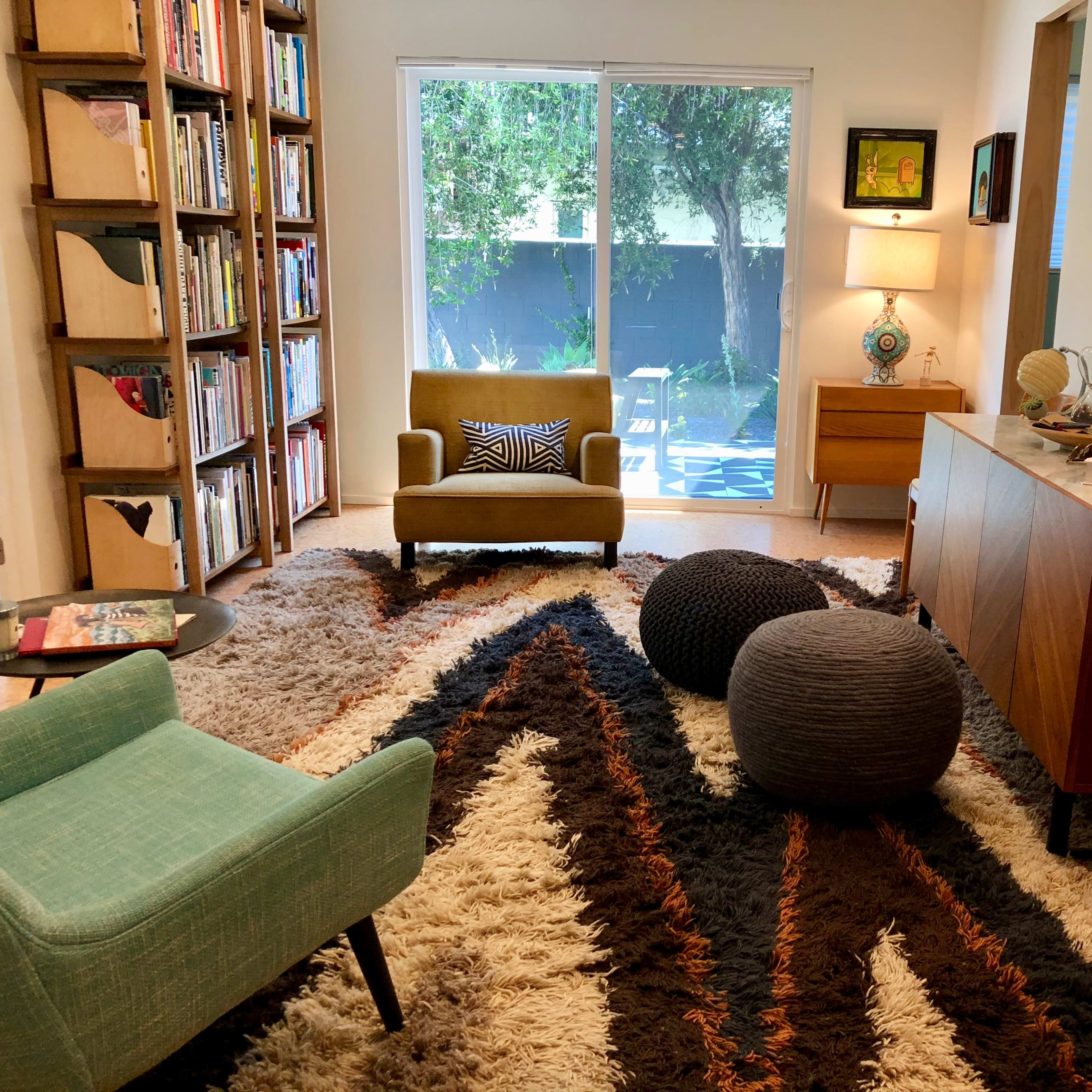 A cozy living room with a bookshelf, armchair, and a patterned rug, featuring a large window that opens to a garden view.