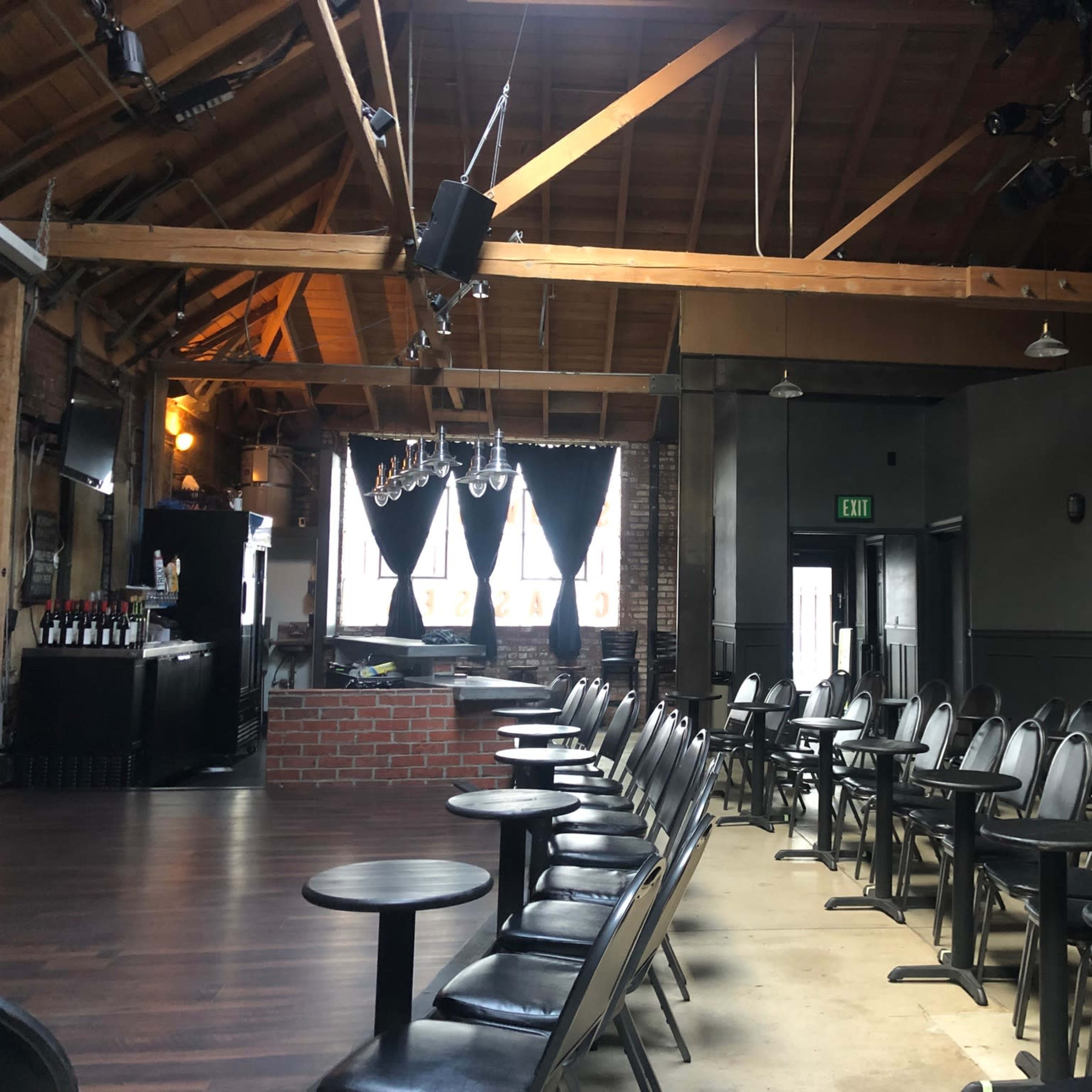 A spacious venue with black chairs and small tables arranged in front of a bar area beneath a wooden ceiling.