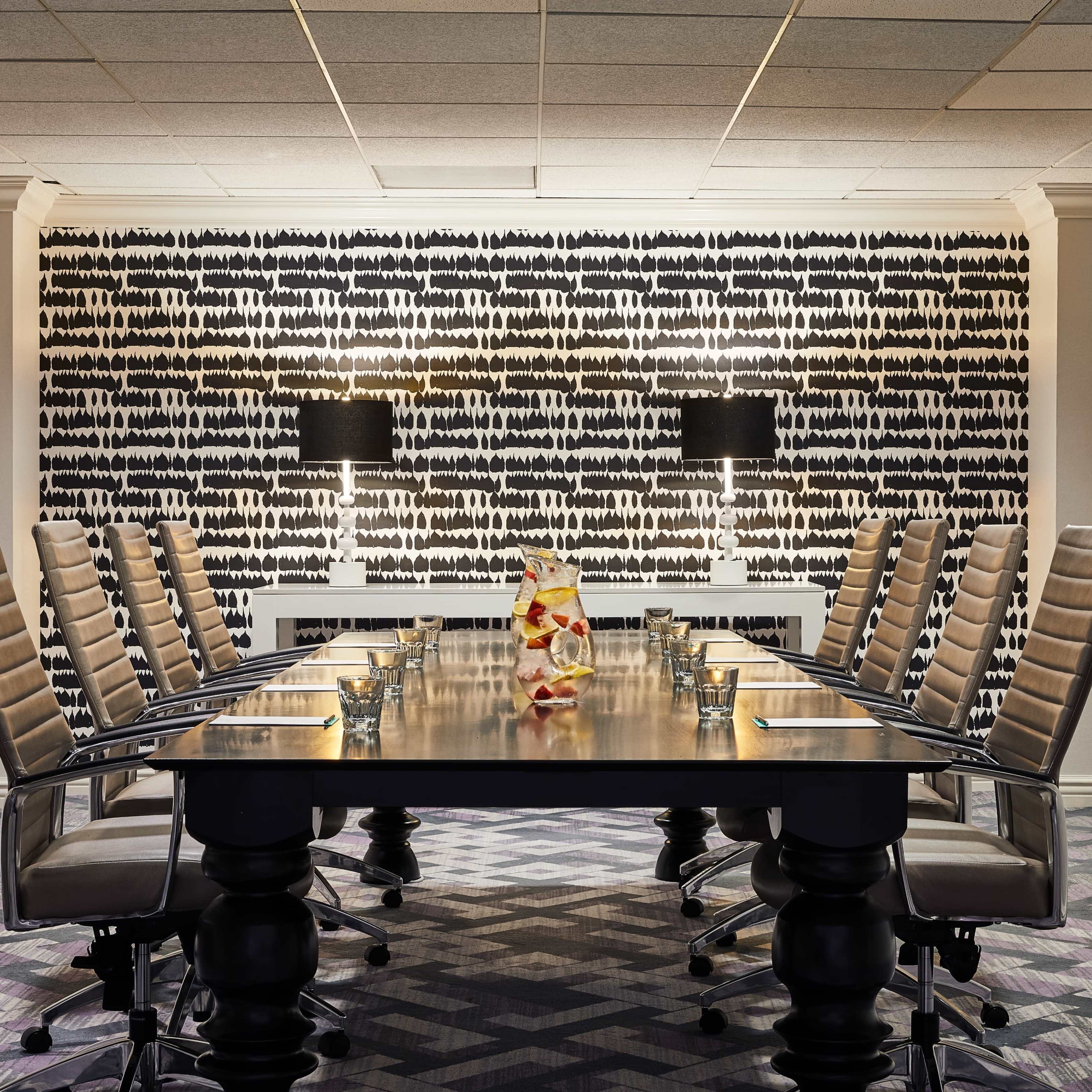 A modern conference room features a long table surrounded by high-backed chairs, with a patterned wall and two lamps providing ambient light.