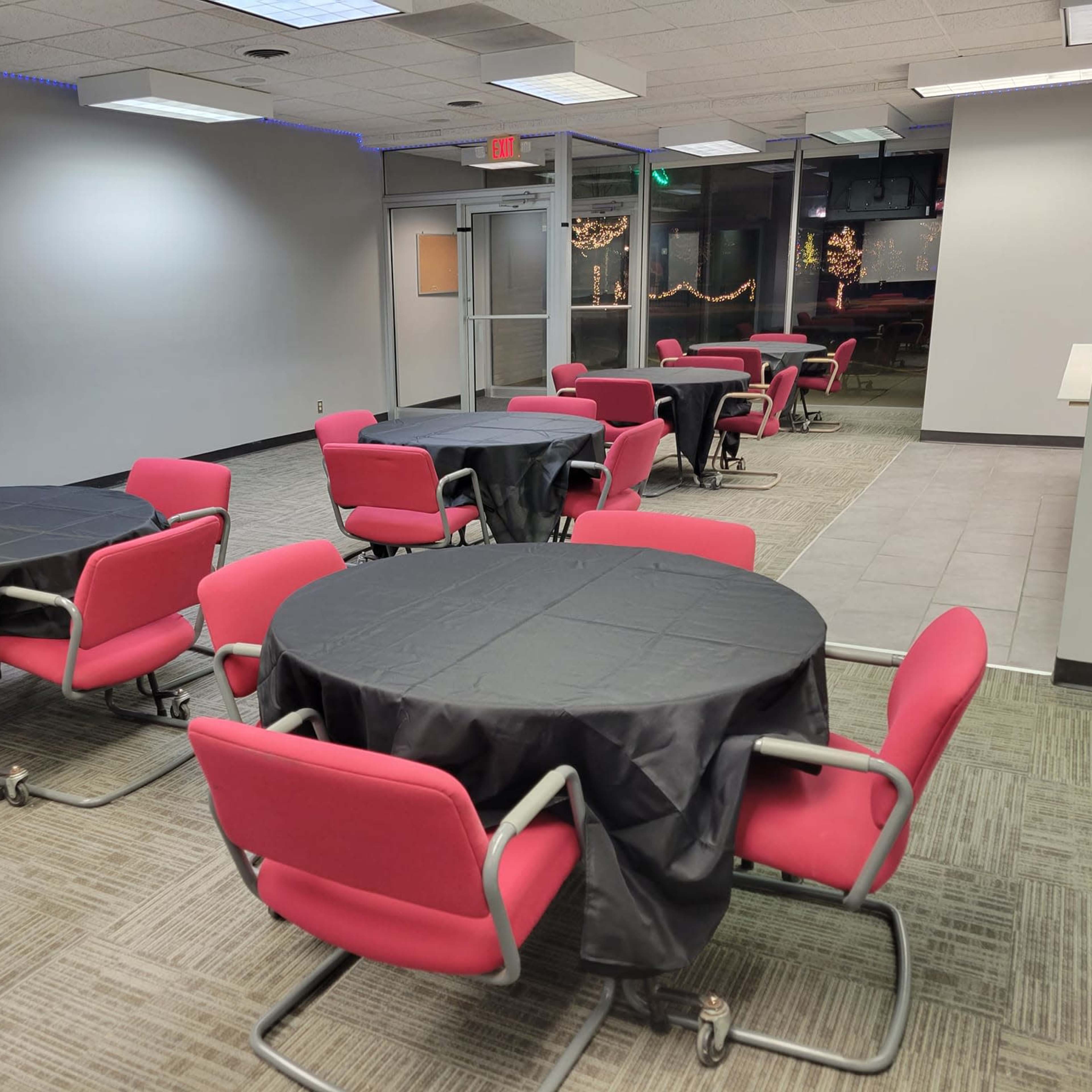 A room with several round tables covered in black tablecloths and surrounded by red chairs, with large windows and a doorway visible at the back.