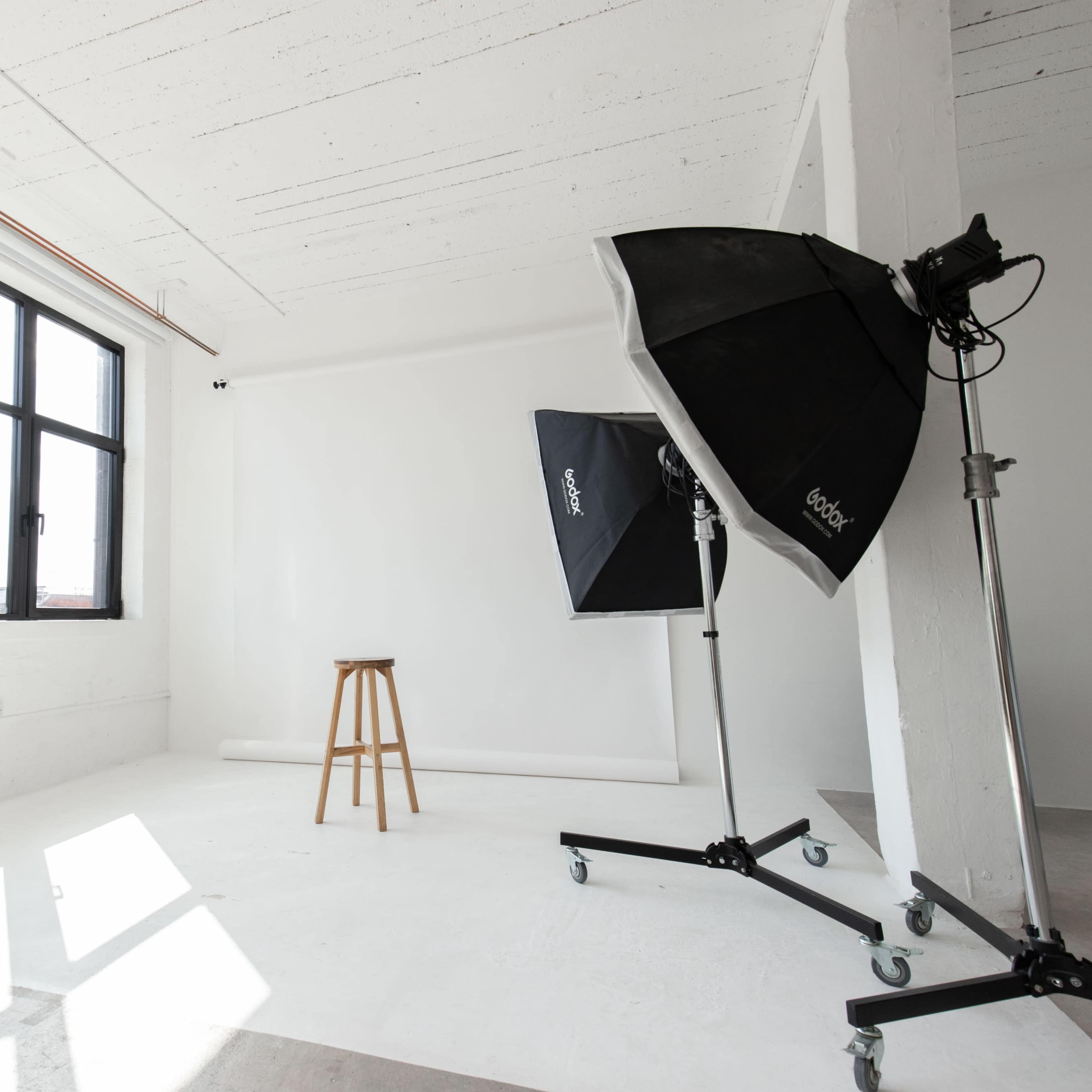 A photography studio with a wooden stool, two lighting umbrellas, and large windows allowing natural light.