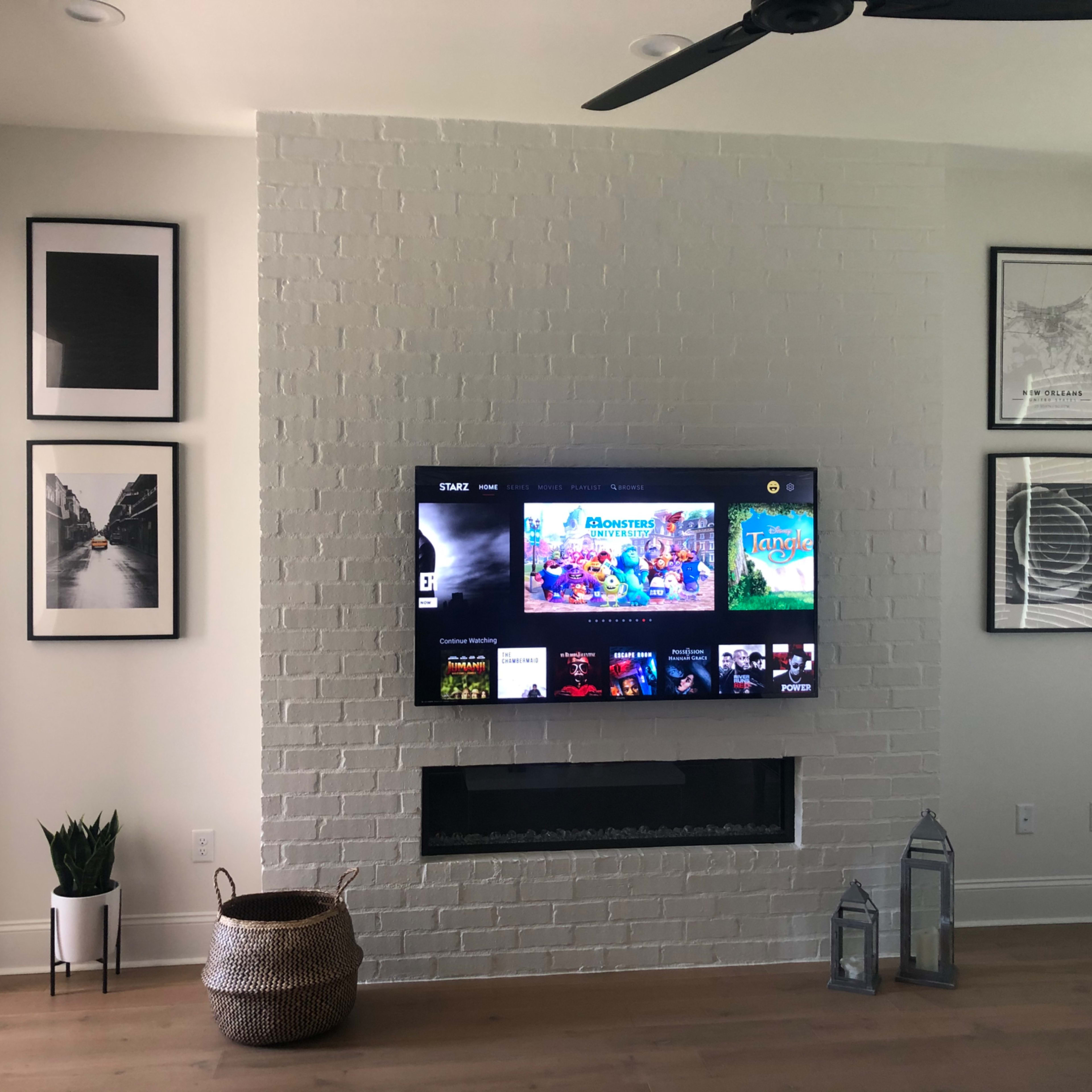 A living room with a white brick wall featuring a mounted television, framed artwork on either side, and a decorative arrangement including a plant and lanterns below the TV.