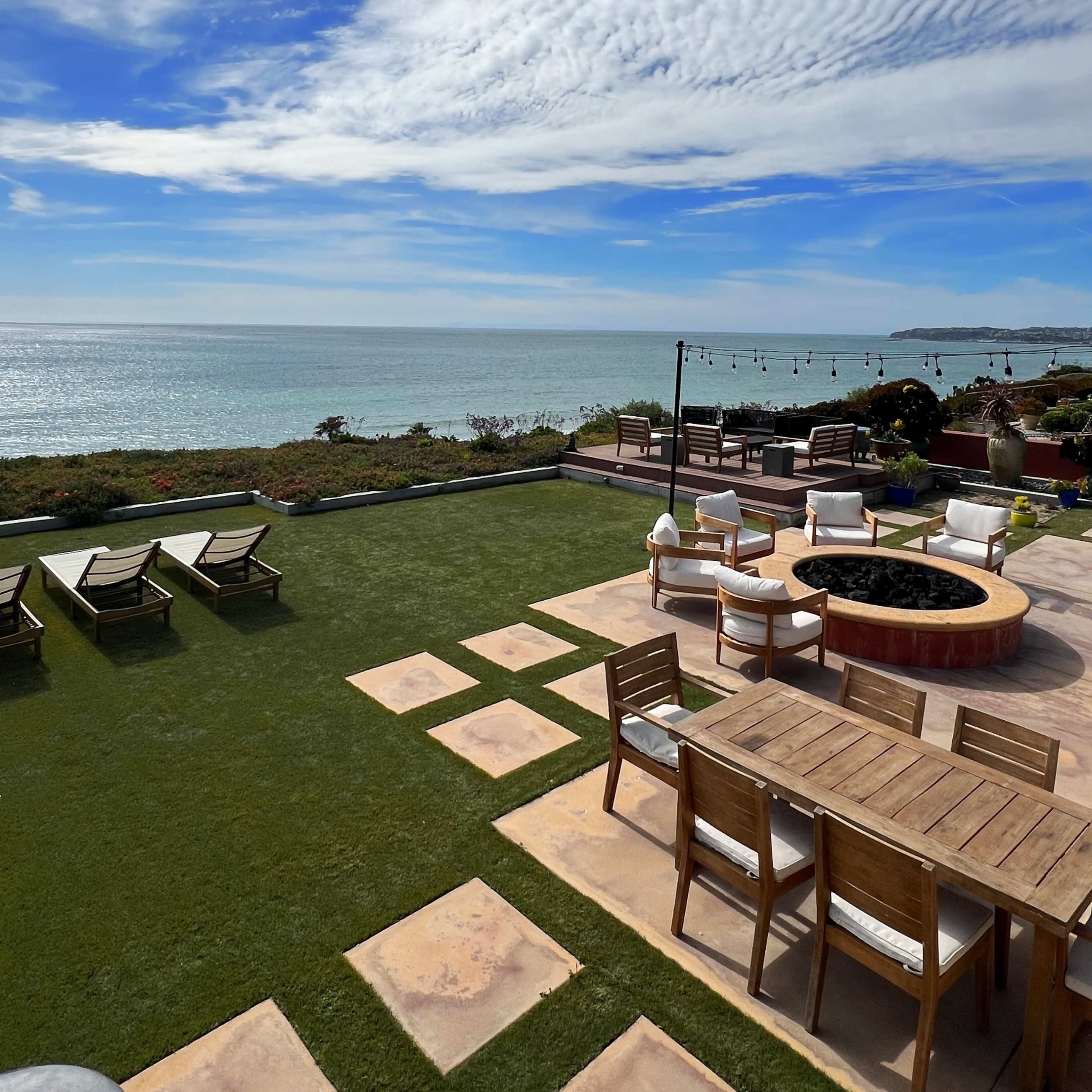 A coastal outdoor space featuring lounge chairs, a fire pit, and a dining table with a view of the ocean and sky.