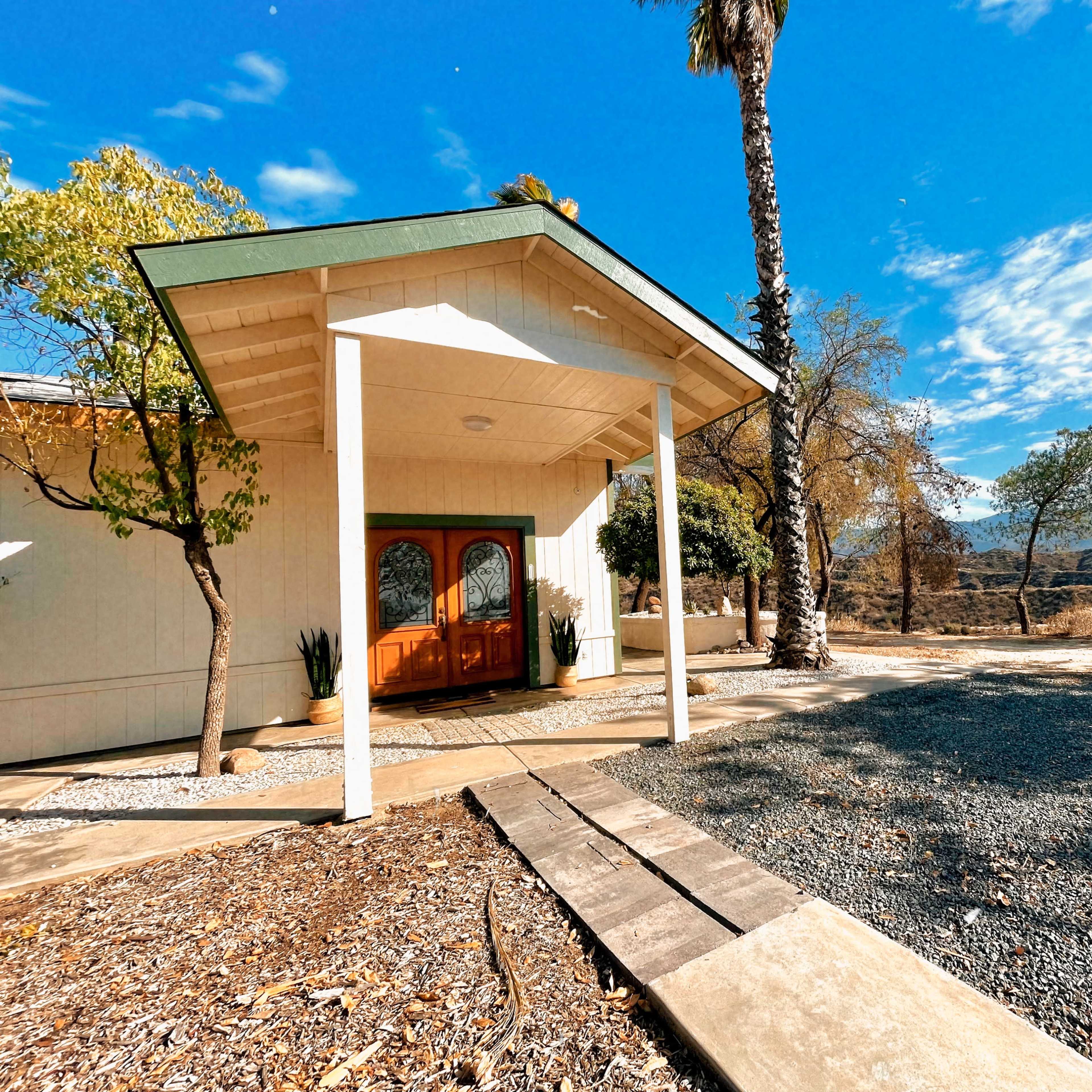 A modern house with a wooden double front door sits under a clear blue sky, surrounded by gravel and sparse trees.