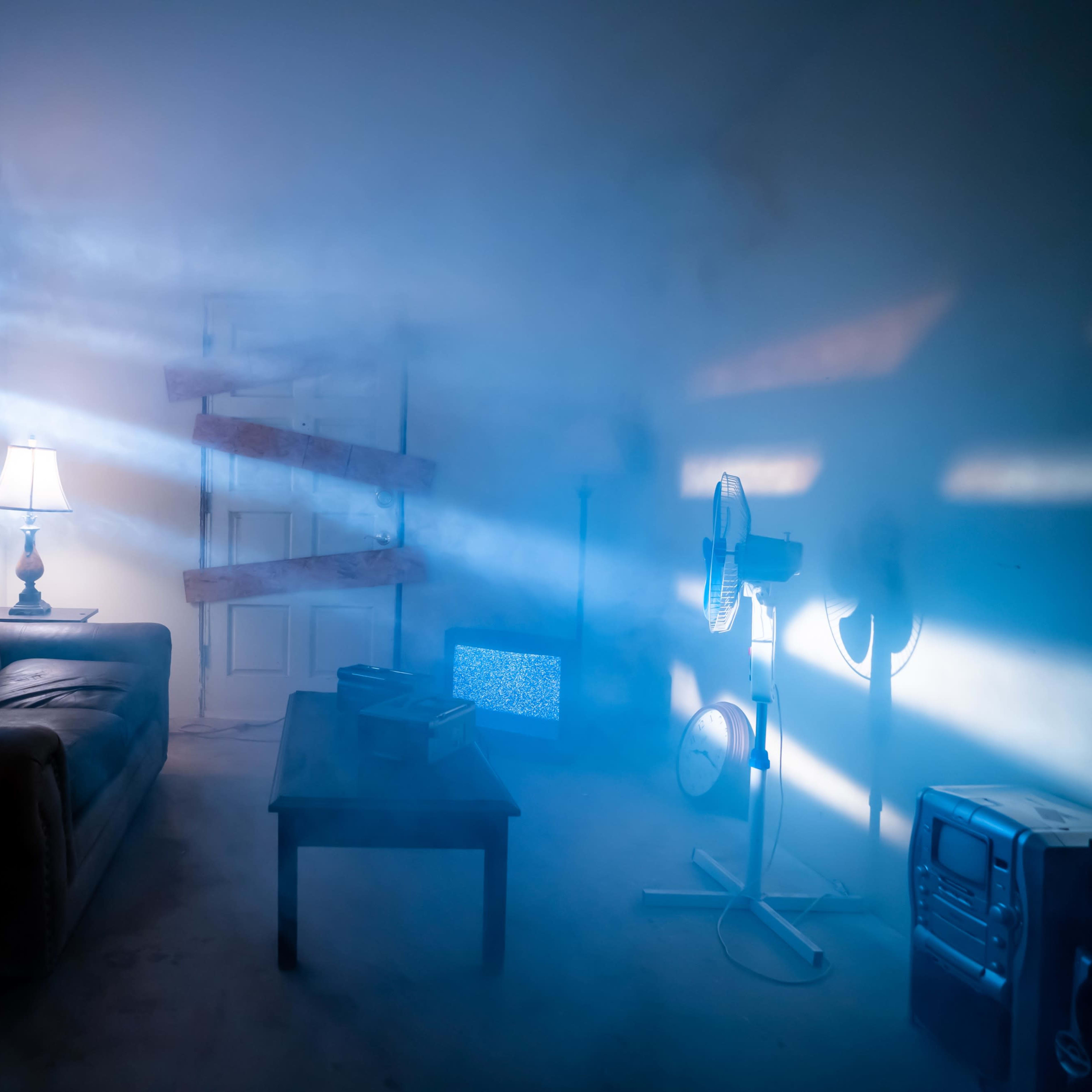 A dimly lit room features a couch, a table, a lamp, and a fan, with beams of light cutting through a mist and a boarded-up door.