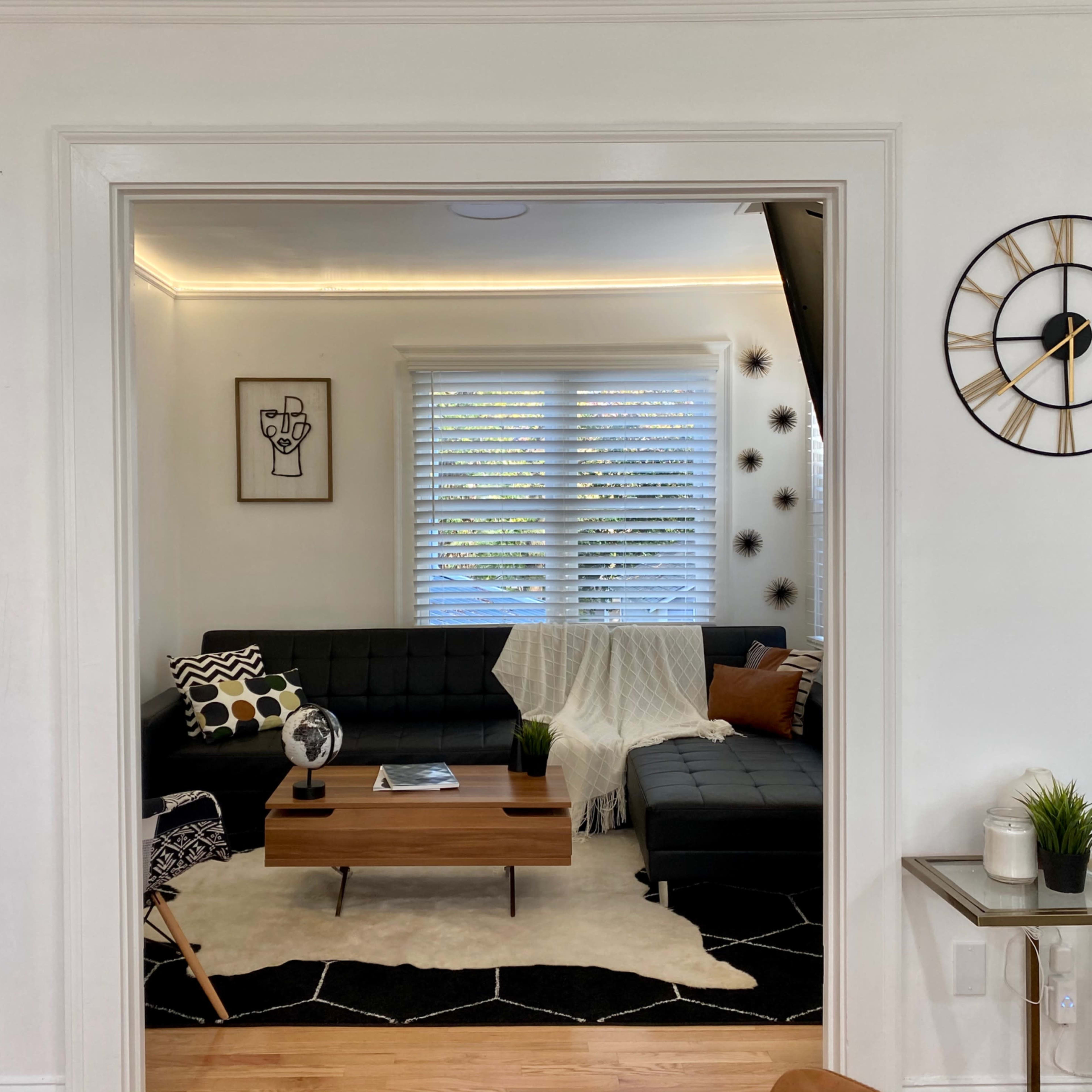 A living room features a dark sofa arrangement, a wooden coffee table, and decorative elements including a plant and a wall clock.