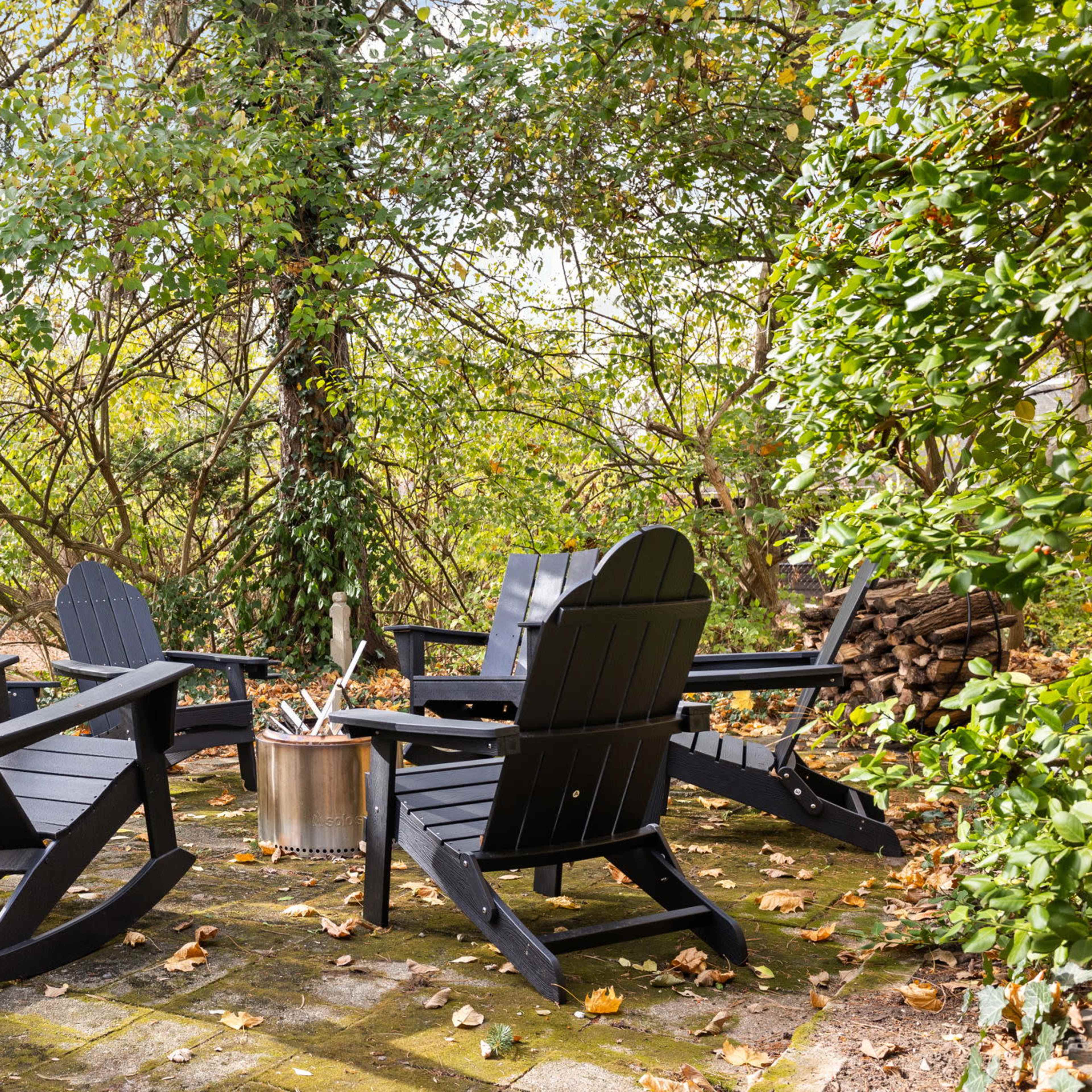 A circular seating area with four black rocking chairs surrounds a metal fire pit, set among greenery and fallen leaves.