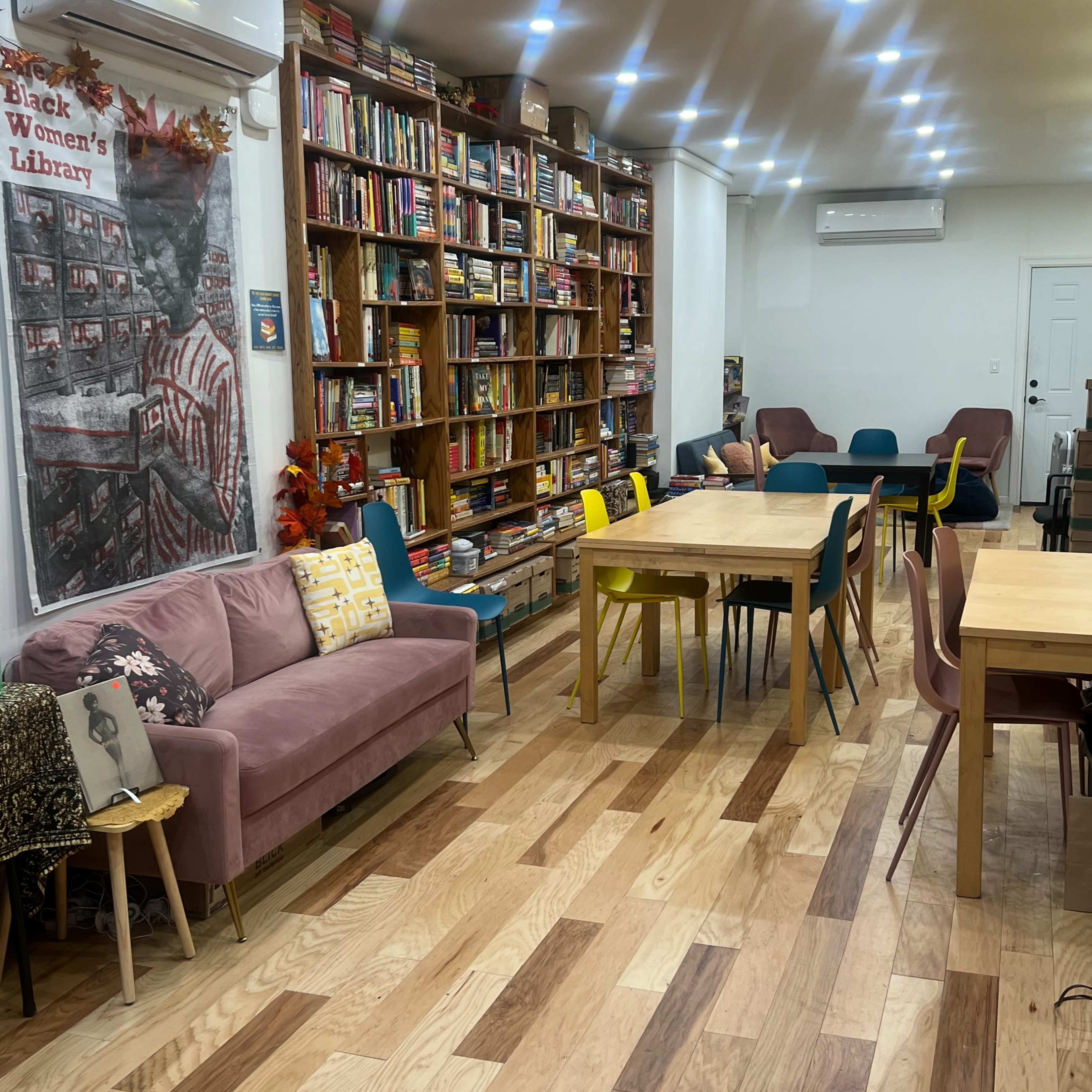 A cozy reading room with a bookshelf filled with books, several tables, and colorful chairs arranged around a wooden floor.