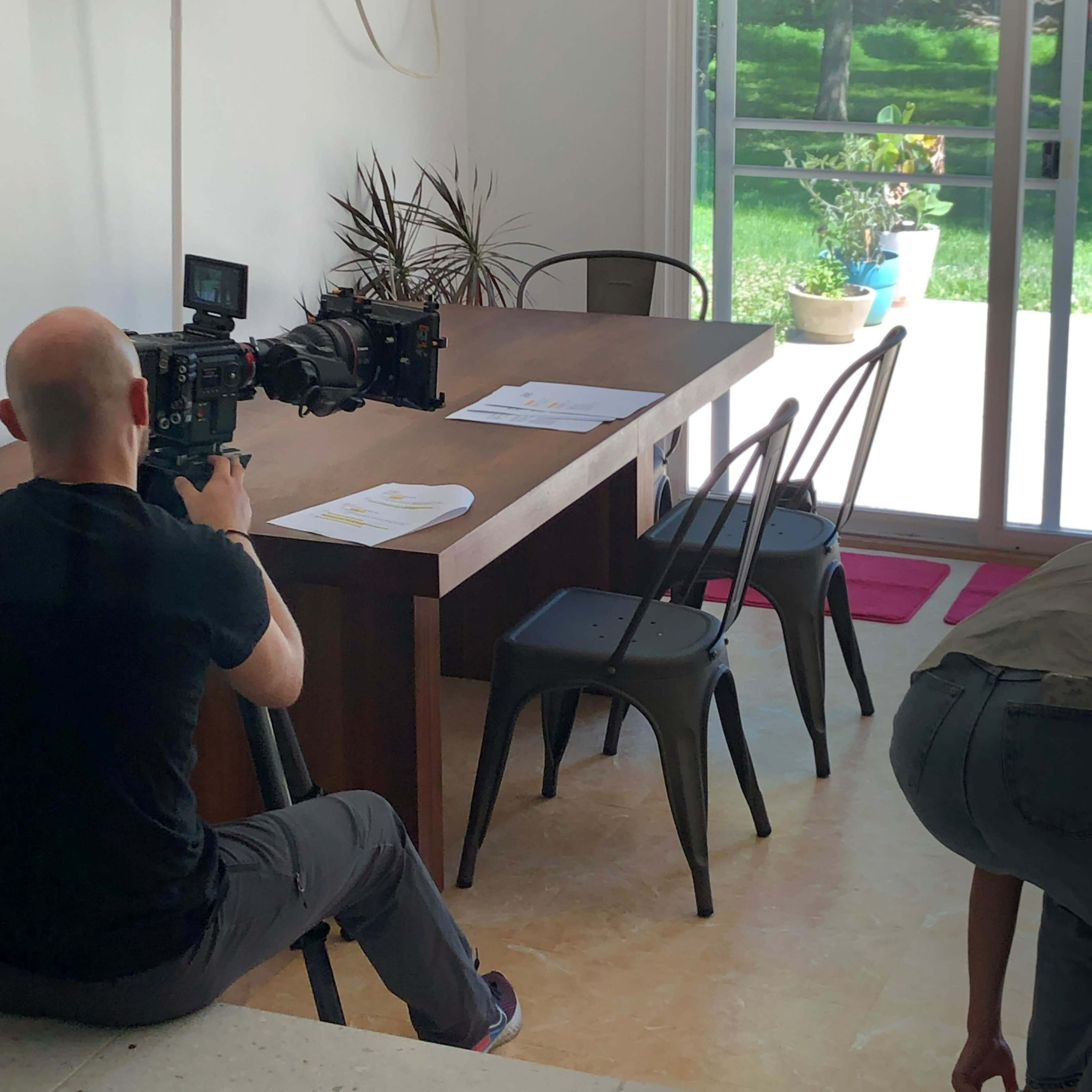 A person is operating a camera on a tripod while filming a scene in a well-lit dining area with a wooden table and several chairs.
