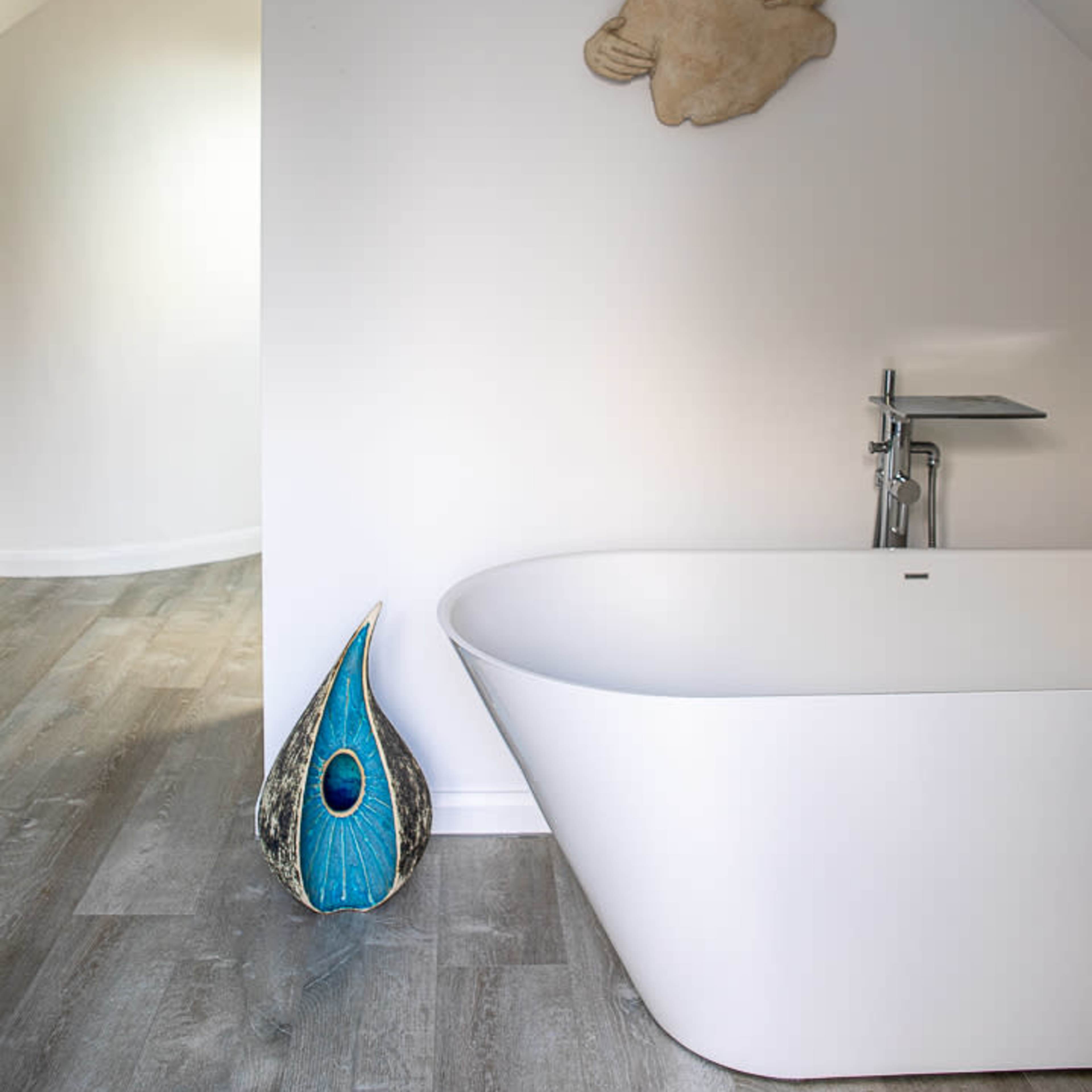 A modern bathroom with a freestanding white bathtub, a decorative blue and black object leaning against the wall, and a textured wall art piece above it.