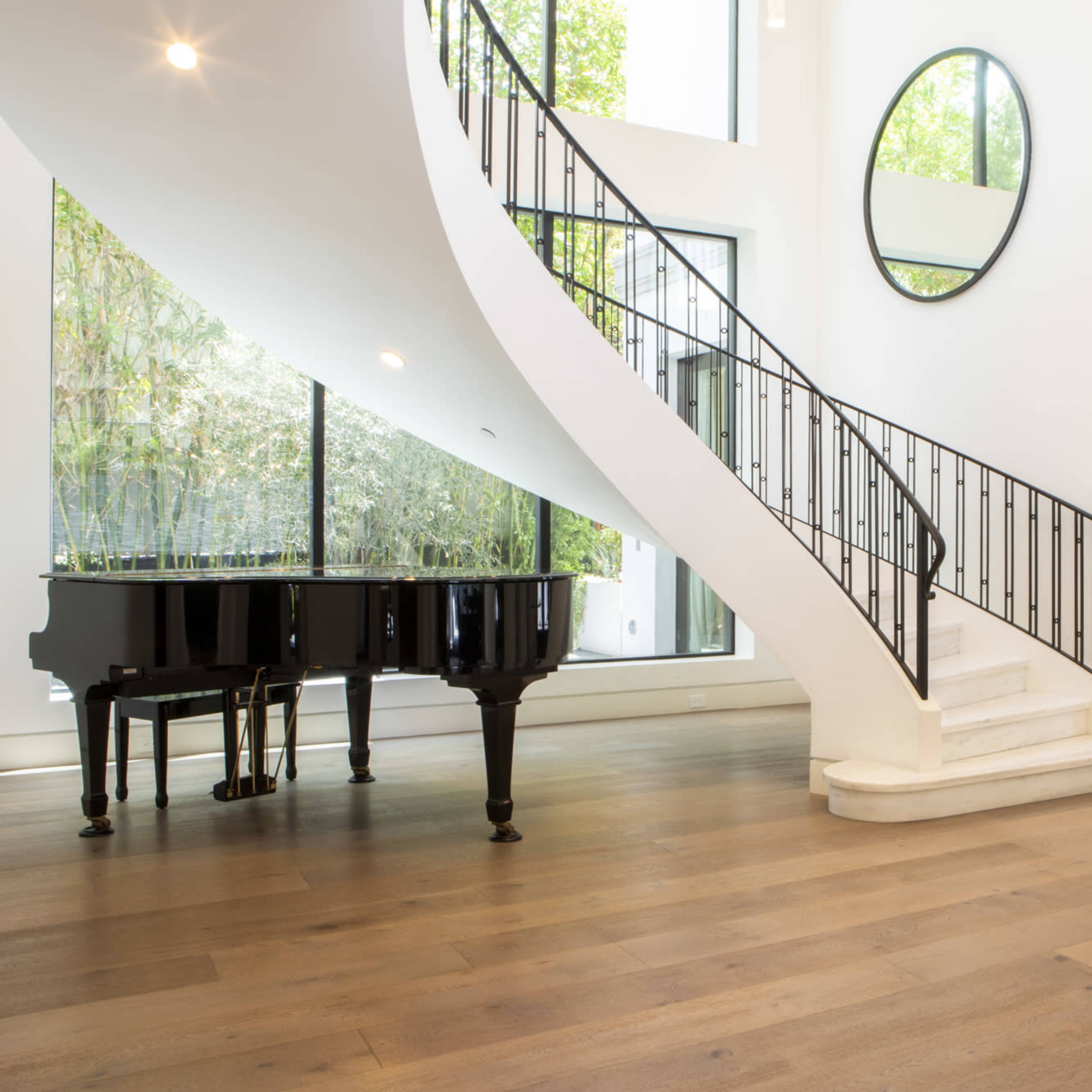 A modern interior featuring a black grand piano next to a curved staircase with a wrought-iron railing and large windows letting in natural light.