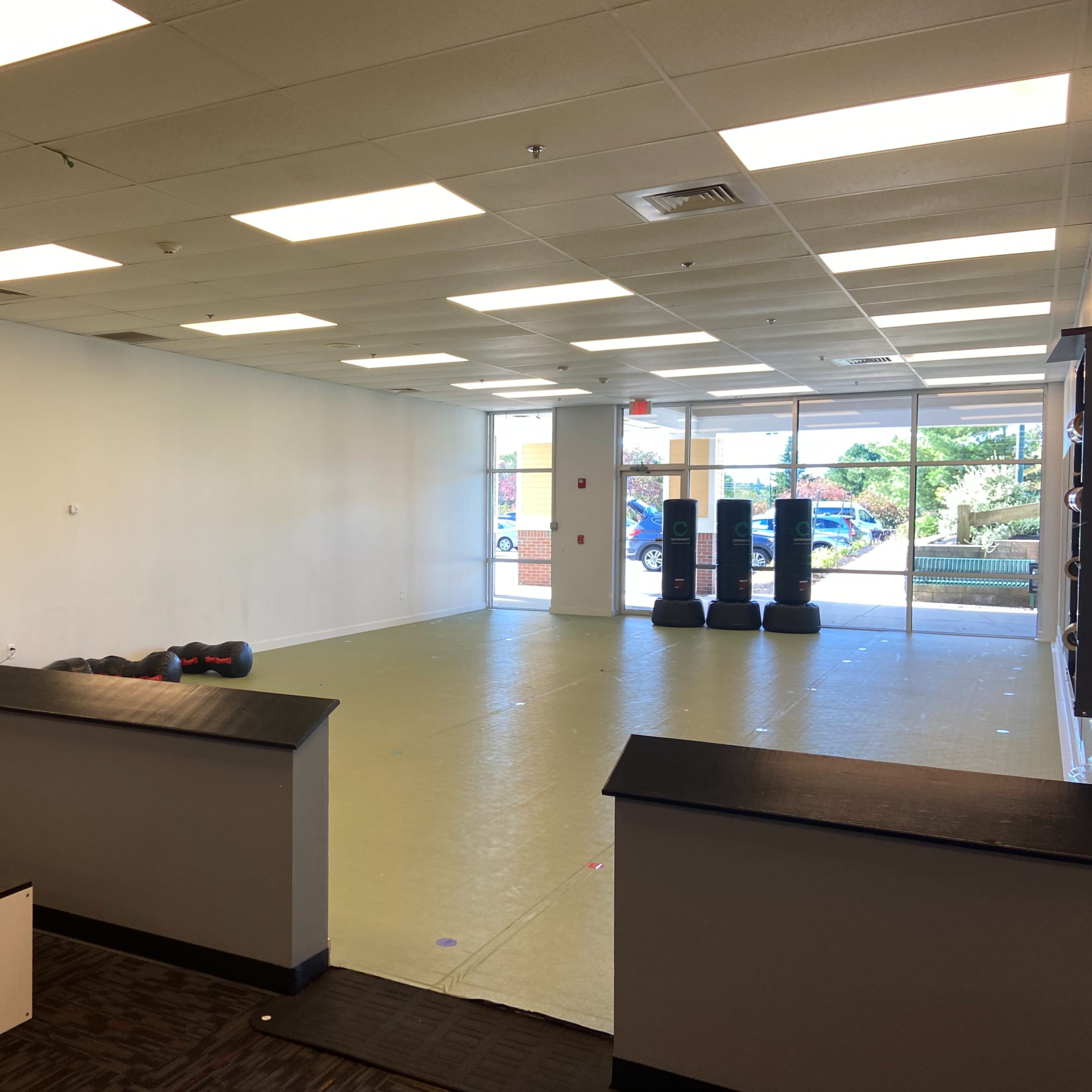 A spacious, empty room with a light-colored floor, large windows, and fitness equipment along one wall.