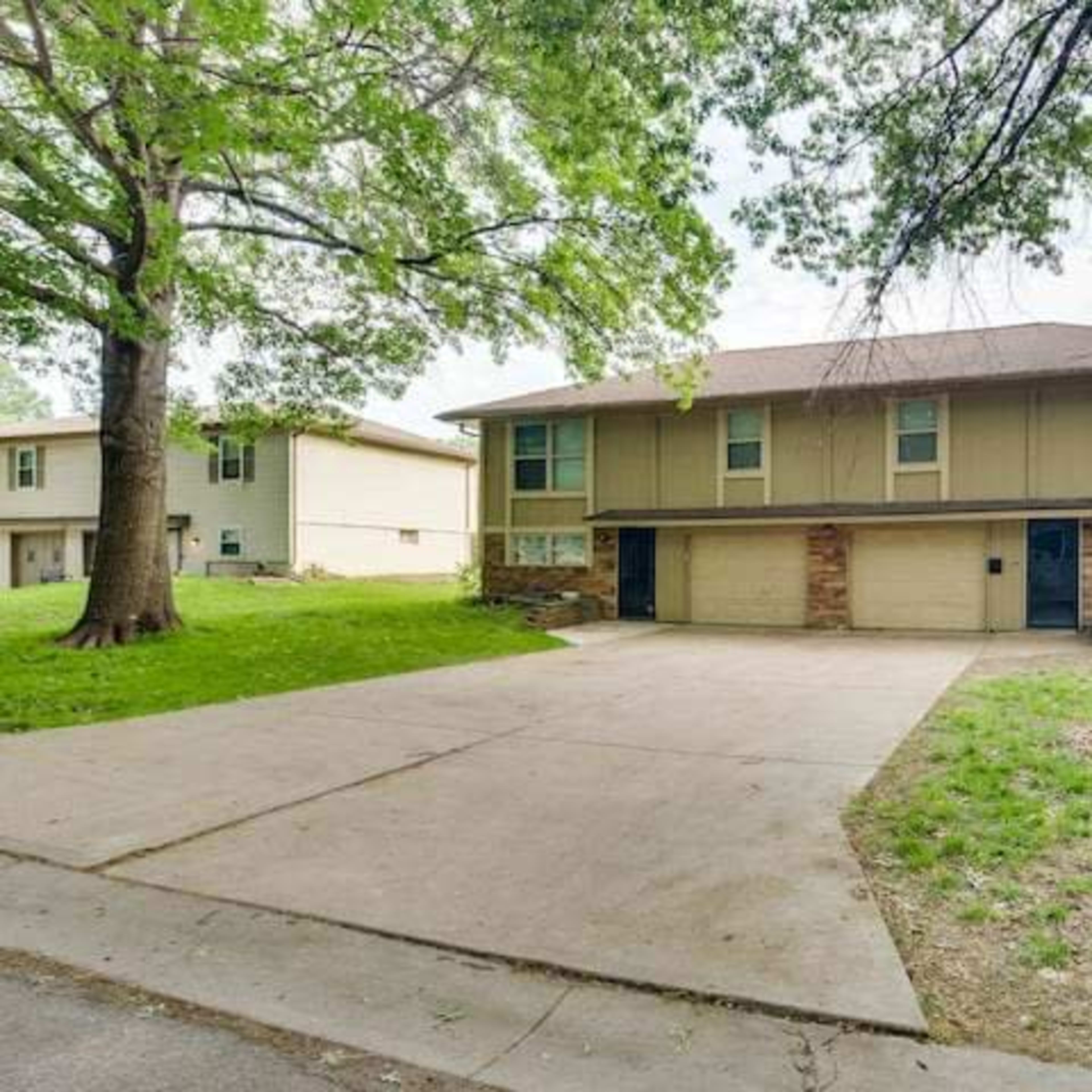 A two-story house with a brick and siding exterior features a double garage and is set on a grassy lot with large trees nearby.