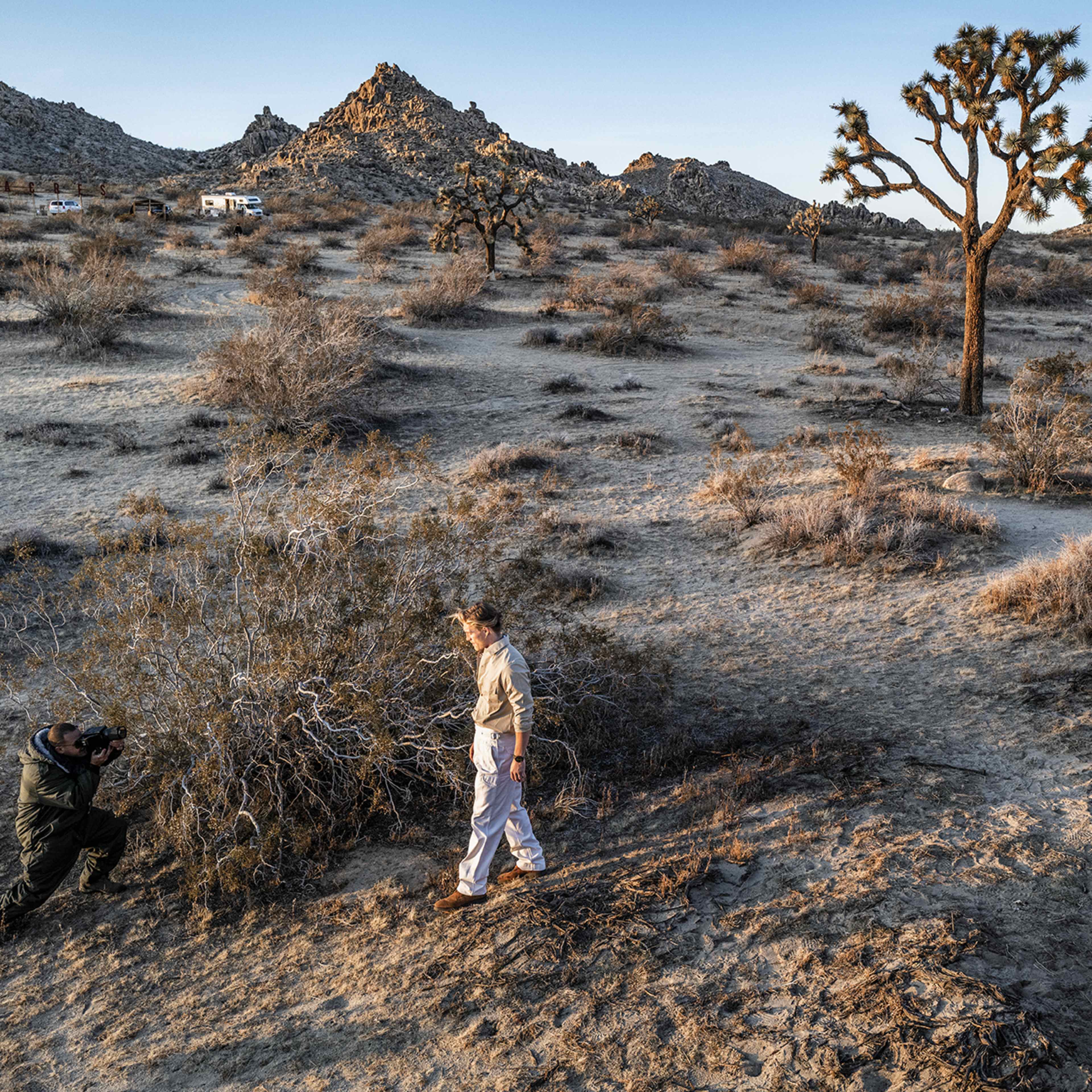 A person poses for a photograph in a desert landscape with scattered shrubs and rocky mountains in the background.