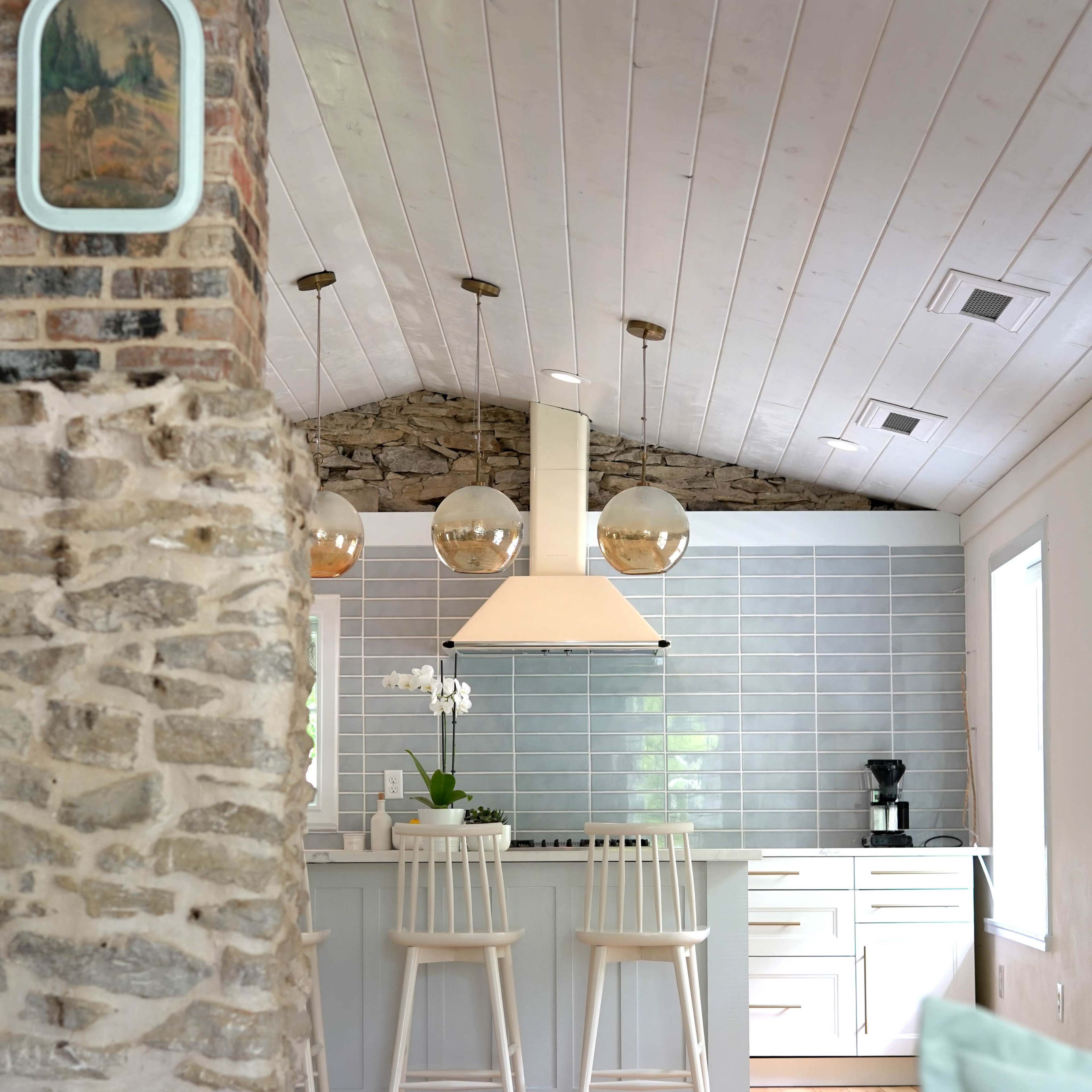 A modern kitchen with a stone pillar, light wood flooring, and a gray tiled backsplash, featuring two white barstools and pendant lights.