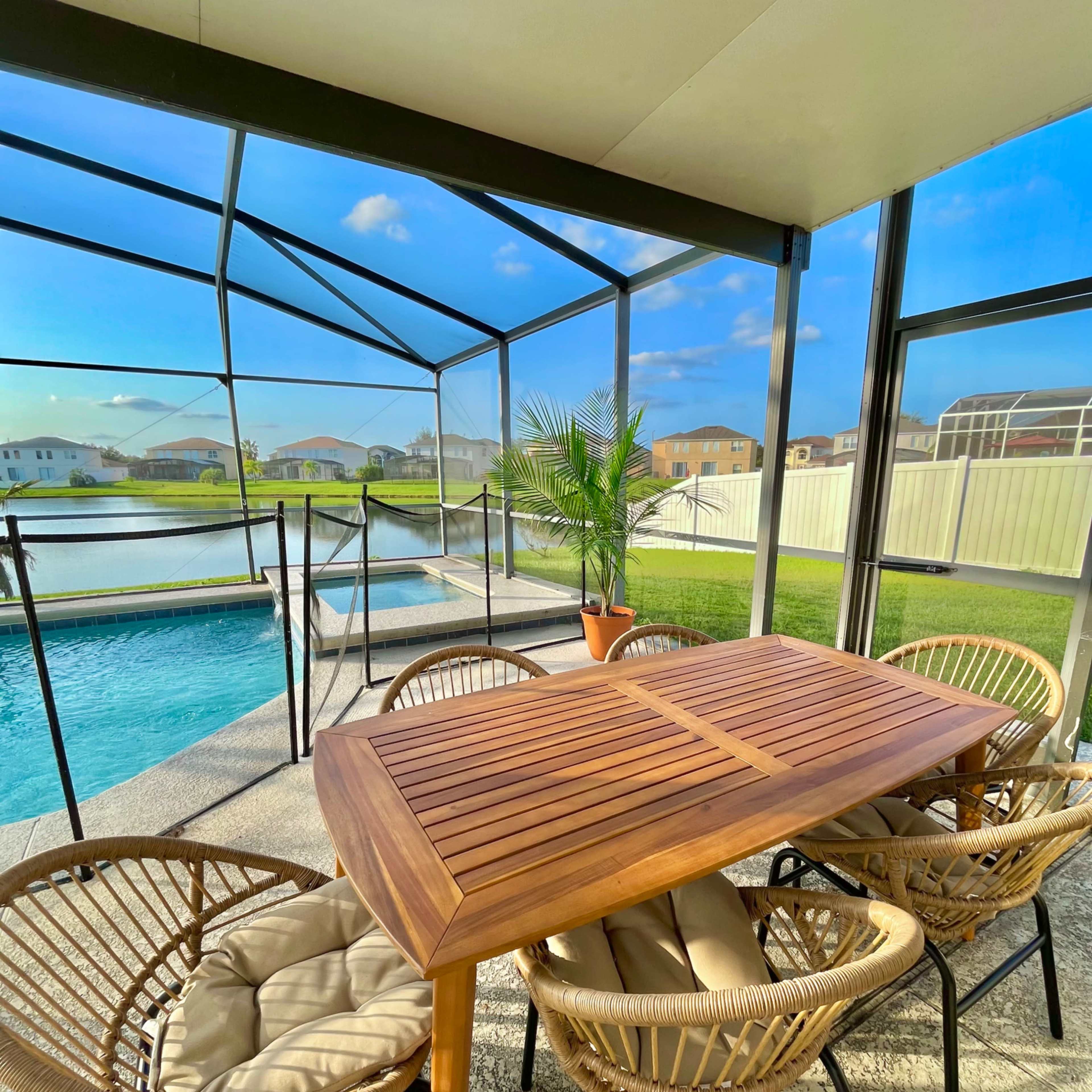 A patio area features a wooden dining table surrounded by wicker chairs, with a pool visible through a screened enclosure.