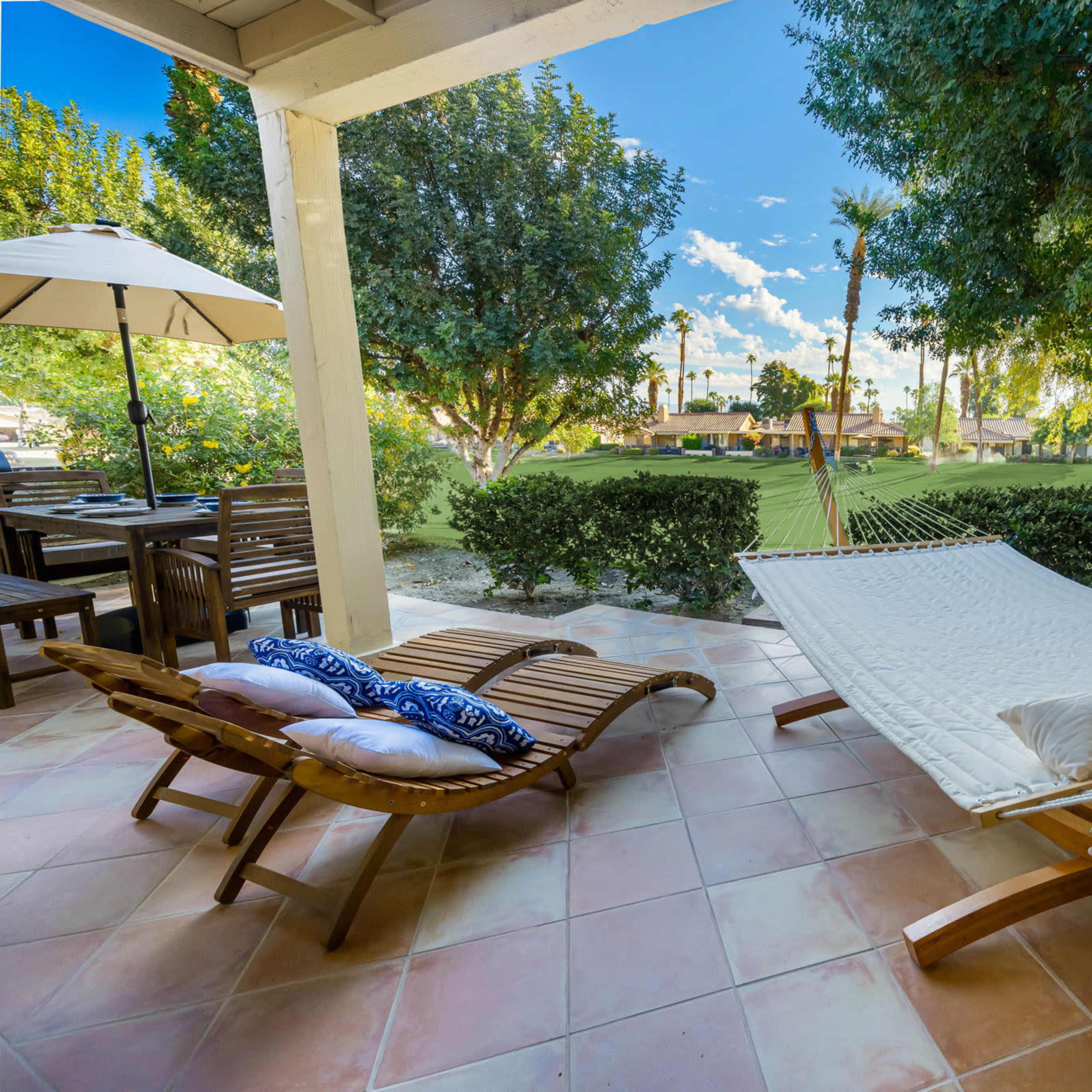 A hammock and a lounge chair are positioned on a patio overlooking a green lawn and trees.