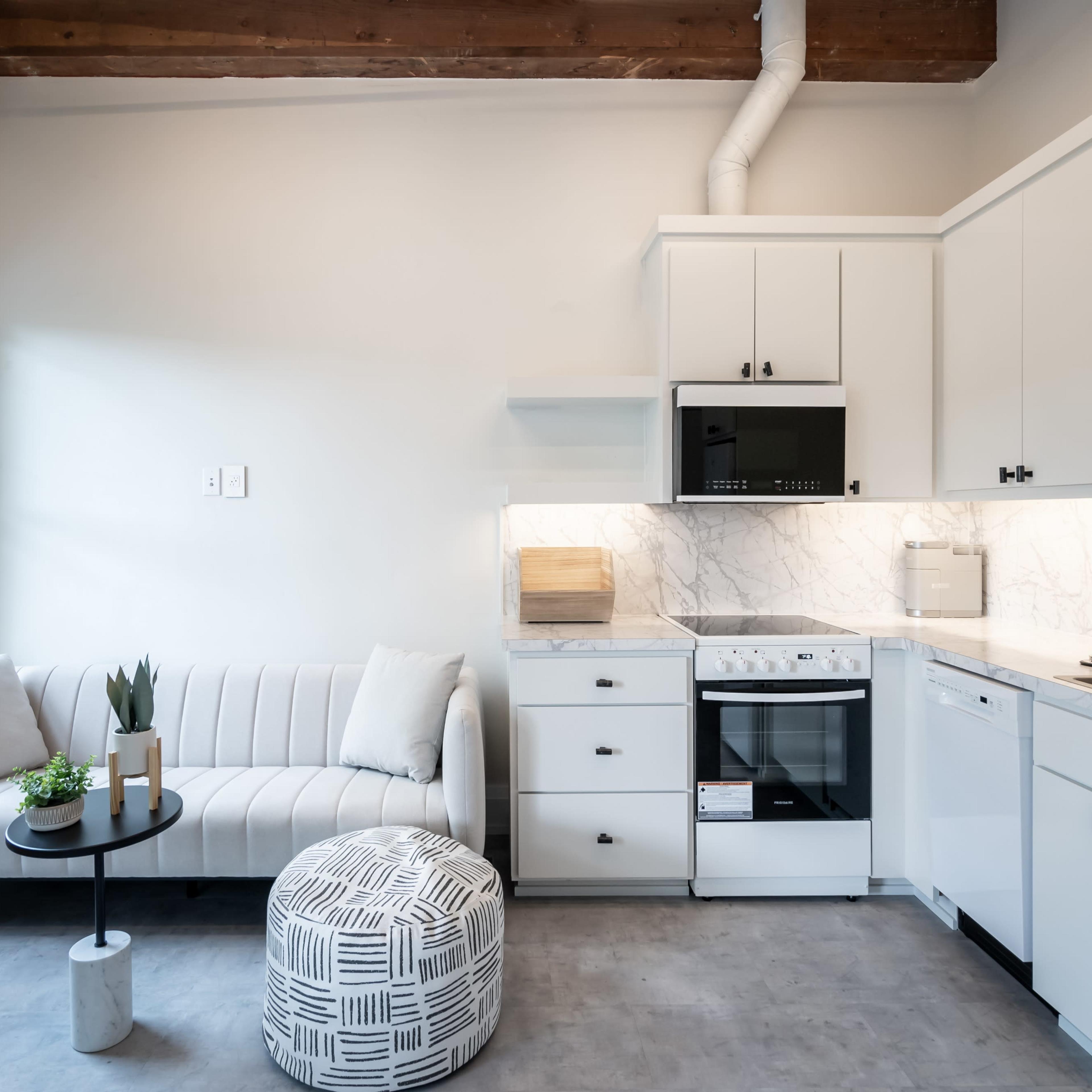 A modern kitchen and living area with a white sofa, a round coffee table, and a compact kitchen featuring light-colored cabinetry and a marble backsplash.