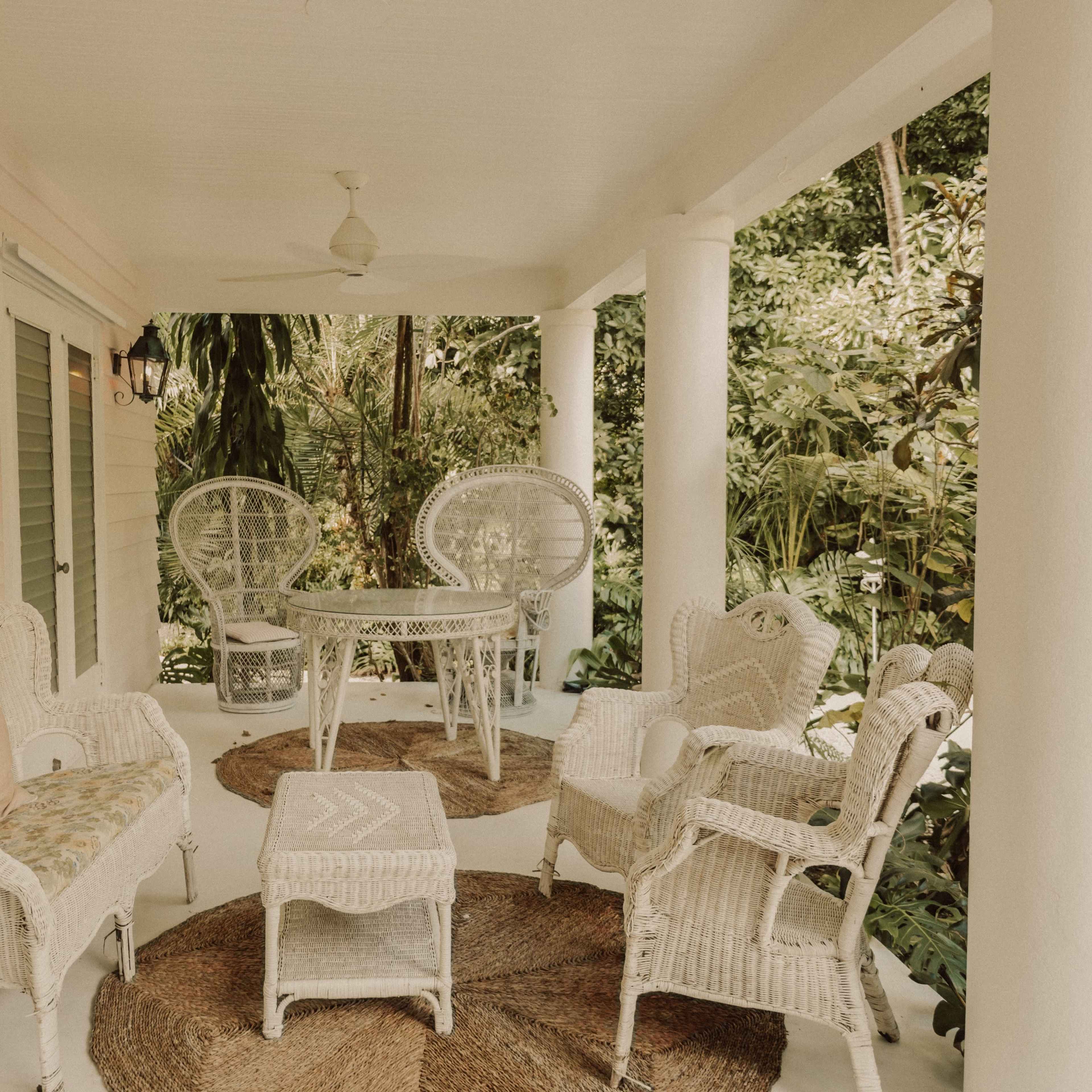 A covered porch features wicker furniture, including chairs and a table, surrounded by tropical greenery.