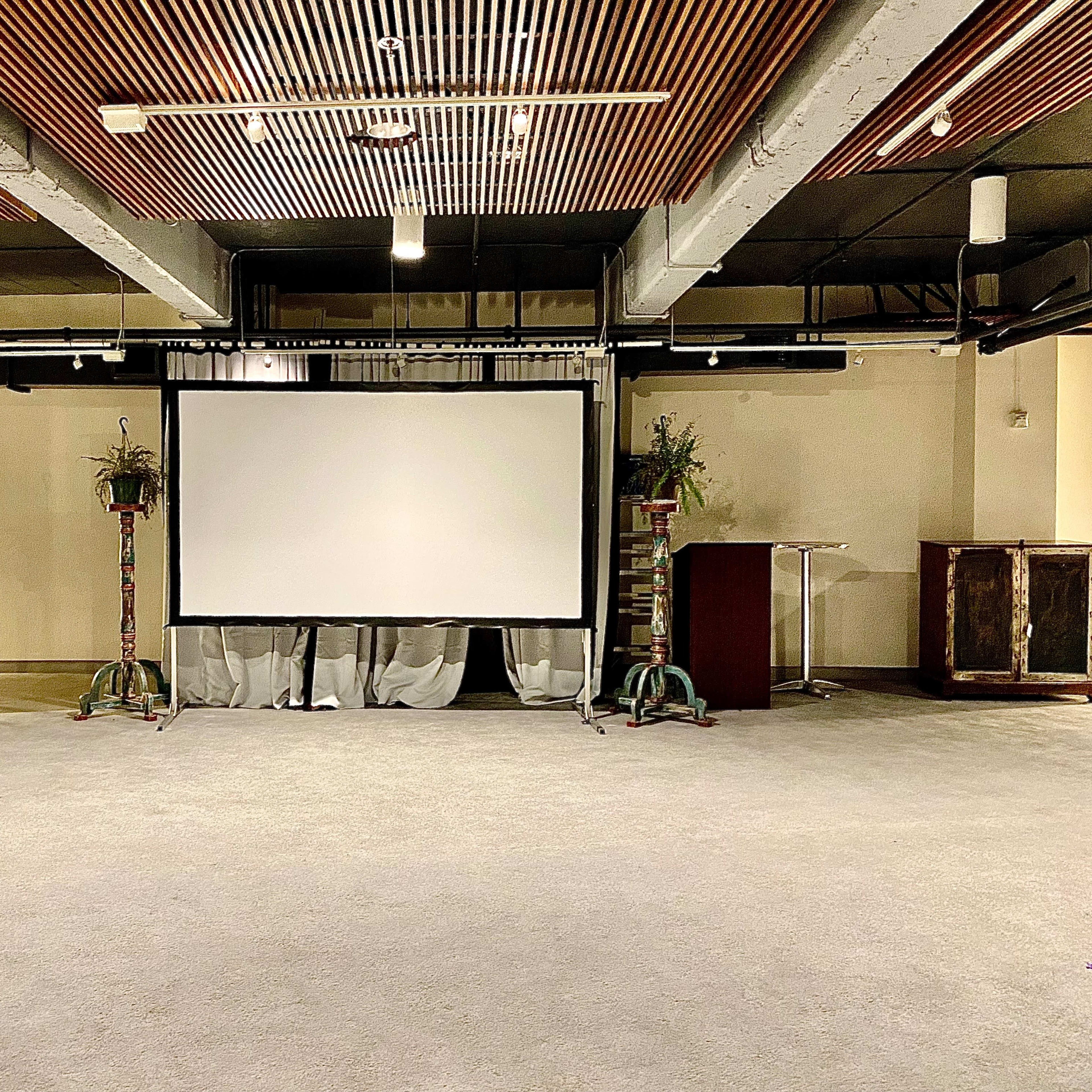 A spacious indoor venue featuring a large projection screen at the center, flanked by decorative plants and furniture.